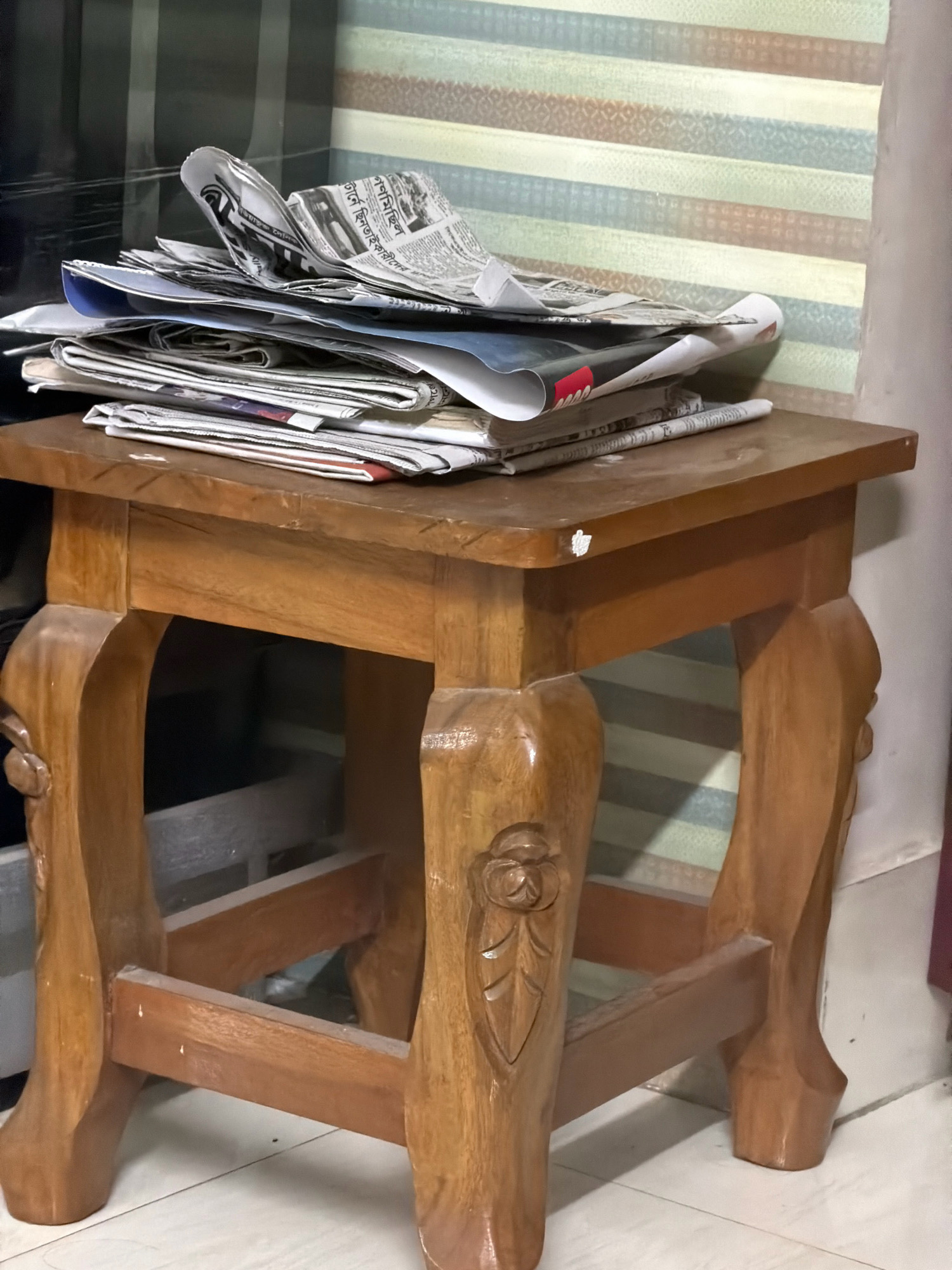 In Bandar, Bangladesh, a wooden stool is positioned on a tiled floor. The stool is made of polished wood and features decorative carvings on its legs. A stack of newspapers is piled on top of the stool. Behind the stool, a striped wallpaper is visible on the wall. The scene appears to be indoors.