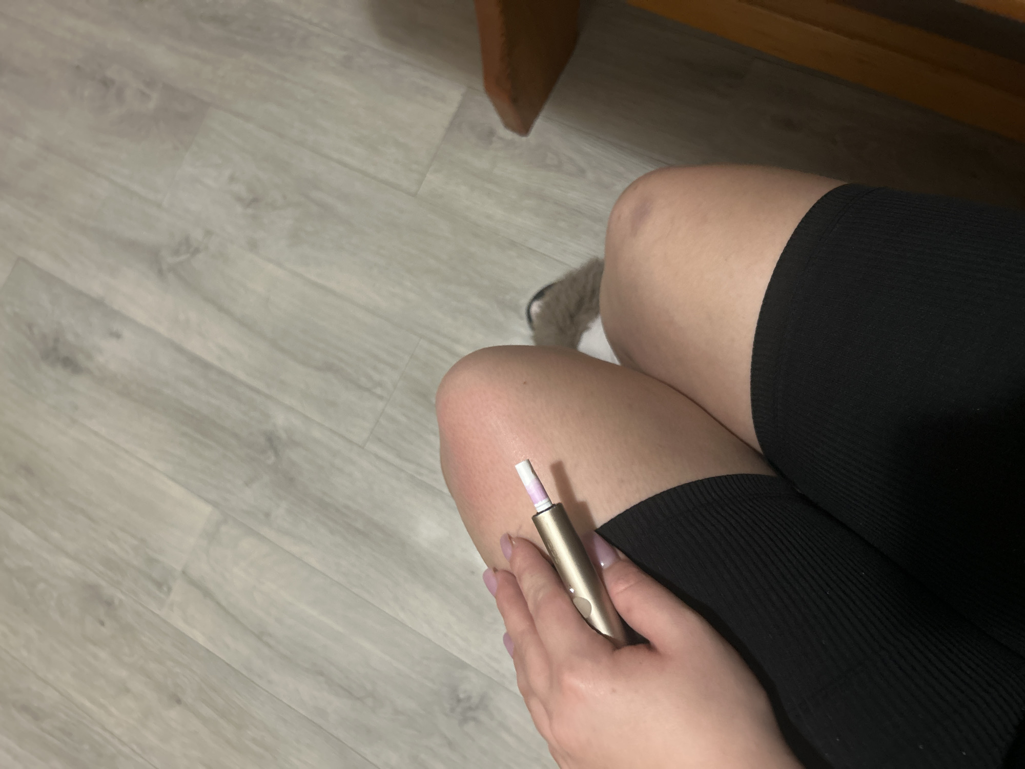 A person's hand, with pale pink nail polish, is holding a gold electronic smoking device near their knee. The person is wearing black, ribbed thigh-high socks. The background shows a light-colored laminate floor. A wooden chair is partially visible on the right side of the image. The image does not contain any information to suggest a location in Basarabeasca, Moldova.