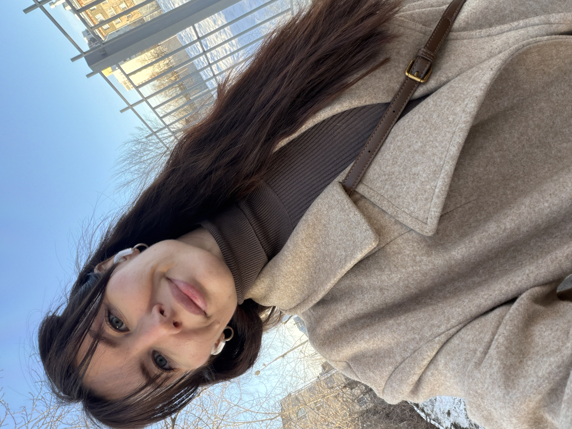 A female subject is depicted in a selfie-style portrait, positioned outdoors under a clear blue sky. The woman has long, dark brown hair with bangs, light skin, and blue-green eyes, exhibiting a gentle smile. She wears a dark brown ribbed turtleneck sweater beneath a light beige, possibly wool-blend, coat. White wireless earbuds are visible in both ears, and a small silver hoop earring is present in her left ear. A brown leather strap with a brass-colored buckle, consistent with a bag strap, traverses her chest.
The background shows an urban environment, with patches of snow on the ground, suggesting a winter or early spring setting. A light-colored metal fence, constructed with vertical and horizontal bars, runs across the mid-ground to the right. Behind the fence and on the left, leafless deciduous trees are visible. In the distant background, multi-story residential buildings of a common architectural style for Russian cities are present. One building appears light yellow or beige. The lighting indicates direct sunlight. While specified as Perm, Russia, the precise location within the city is indeterminate from the visual information alone due to the generic nature of the background elements.