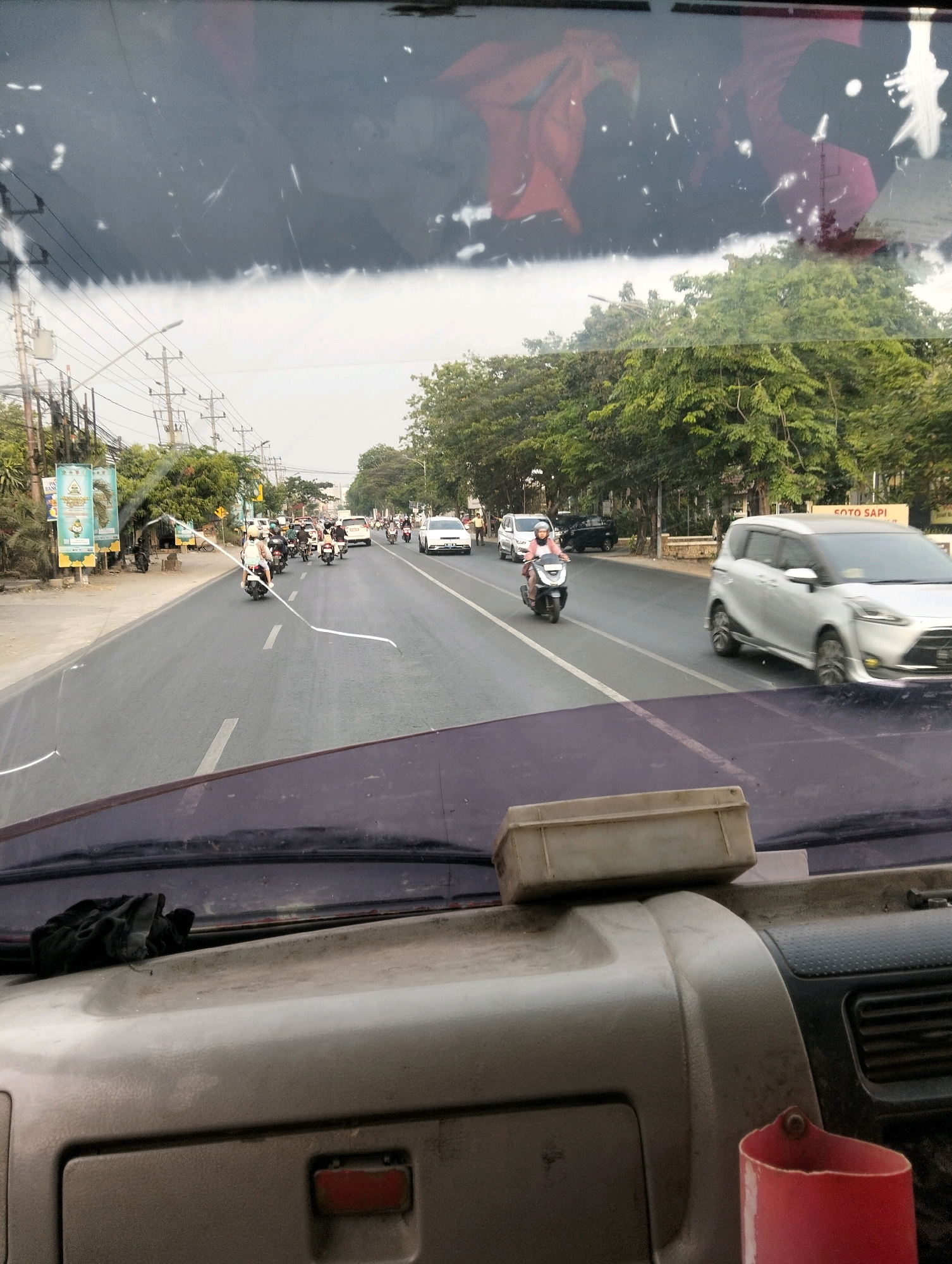 The image depicts a view from inside a vehicle, looking out through the windshield onto a busy street in Semarang, Indonesia. The foreground shows the dashboard of the vehicle, with a beige box resting on top. The windshield itself is dusty and shows some reflections.
The street scene outside is characterized by moderate traffic. Several motorcycles, a common mode of transport in Indonesia, are visible, some carrying single riders and others with passengers. A few cars are also present, including a silver minivan and a white sedan. The road appears to be paved and has lane markings.
On the left side of the road, there are sidewalks, utility poles with numerous wires, and some vegetation. A billboard with Indonesian text and images is also visible. On the right side of the road, there are trees lining the street and buildings in the background, including one with a sign that appears to read "SOTO SAPI" (beef soup), indicating a food establishment. The sky is overcast, suggesting a cloudy day. The overall impression is of a typical urban street environment in a Southeast Asian city during daylight hours.