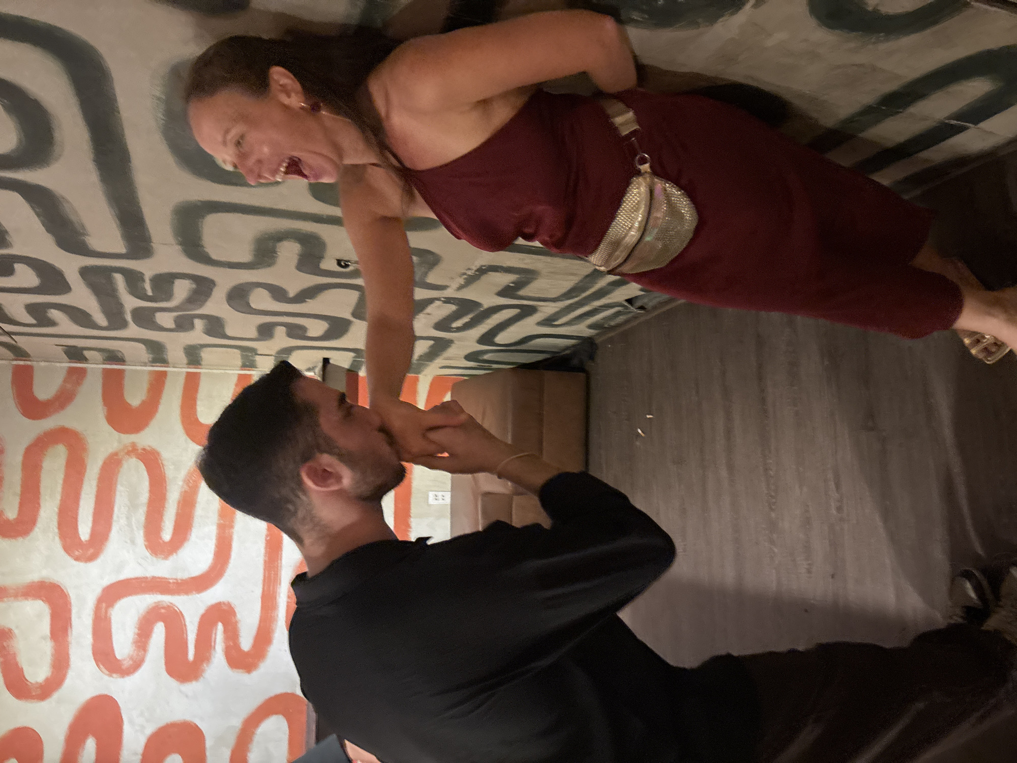A man in a black long-sleeved shirt and dark pants kisses a woman's hand. The woman, wearing a burgundy dress and a shimmering gold fanny pack, leans against a wall and laughs. The wall behind them is decorated with abstract, winding patterns in orange and black. The flooring appears to be made of wooden planks. A tan couch is visible in the background. The overall setting suggests an indoor event or establishment. The city of Đà Nẵng, Vietnam is not identifiable from the image.