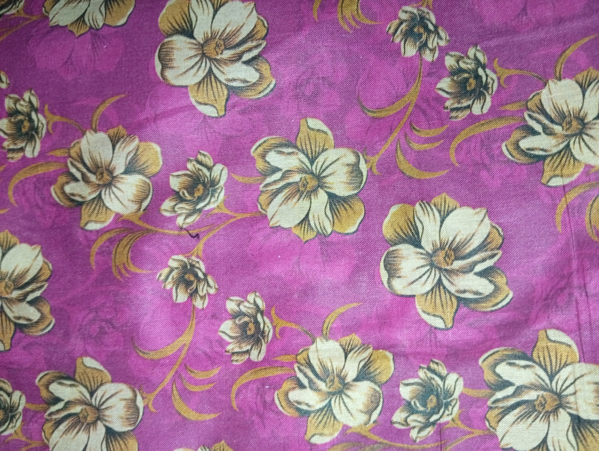 The image shows a close-up of a pink fabric with a floral pattern. The flowers are depicted in shades of cream and gold, with brown outlines. Swirling golden tendrils connect the flowers. The fabric appears to be slightly textured and wrinkled, suggesting it might be a piece of clothing, upholstery, or bedding. The lighting is soft, casting subtle shadows. There is no discernible urban environment or specific location in Larkana, Pakistan, visible in this image. The scene is solely focused on the textile itself.