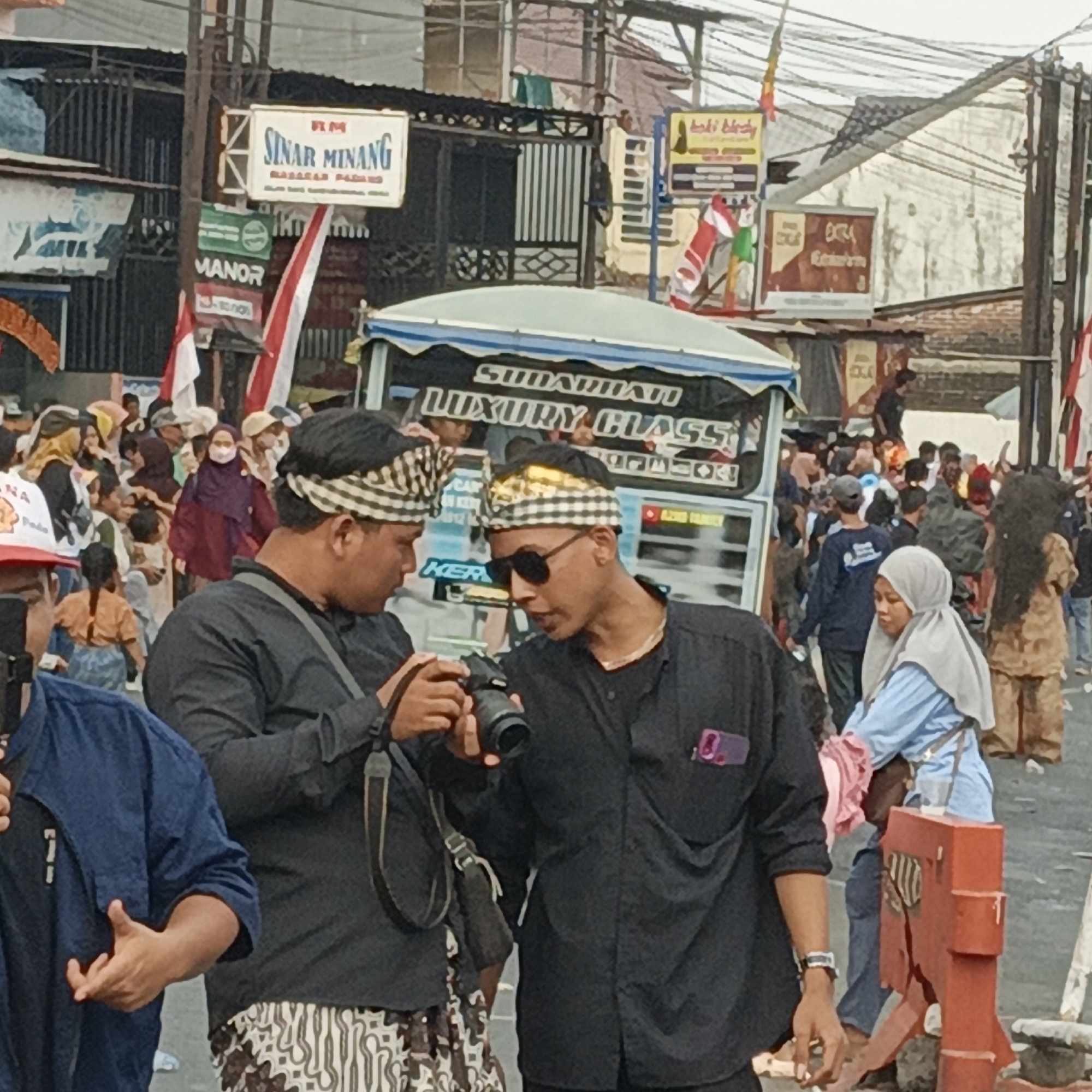 This image captures a bustling street scene in Randudongkal, Indonesia, likely during a local event or festival. In the foreground, two men are engaged in conversation. The man on the left, wearing a black shirt and a patterned head covering, is holding a camera. The man on the right, also in a black shirt and similar head covering, wears sunglasses and appears to be showing something on the camera to his companion. Both men are wearing patterned pants, possibly batik.
Behind them, a crowd of people of various ages is visible, many dressed in traditional attire or casual clothing. Some individuals are wearing face masks. A large truck with the text "SUDAHA MAJU LUXURY CLASS" is prominently displayed, suggesting it might be part of a parade or transportation for an event. Several Indonesian flags are displayed, adding to the festive atmosphere. Buildings and various signs are visible in the background, indicative of a commercial or residential area. The overall environment suggests an outdoor gathering with a lively ambiance.