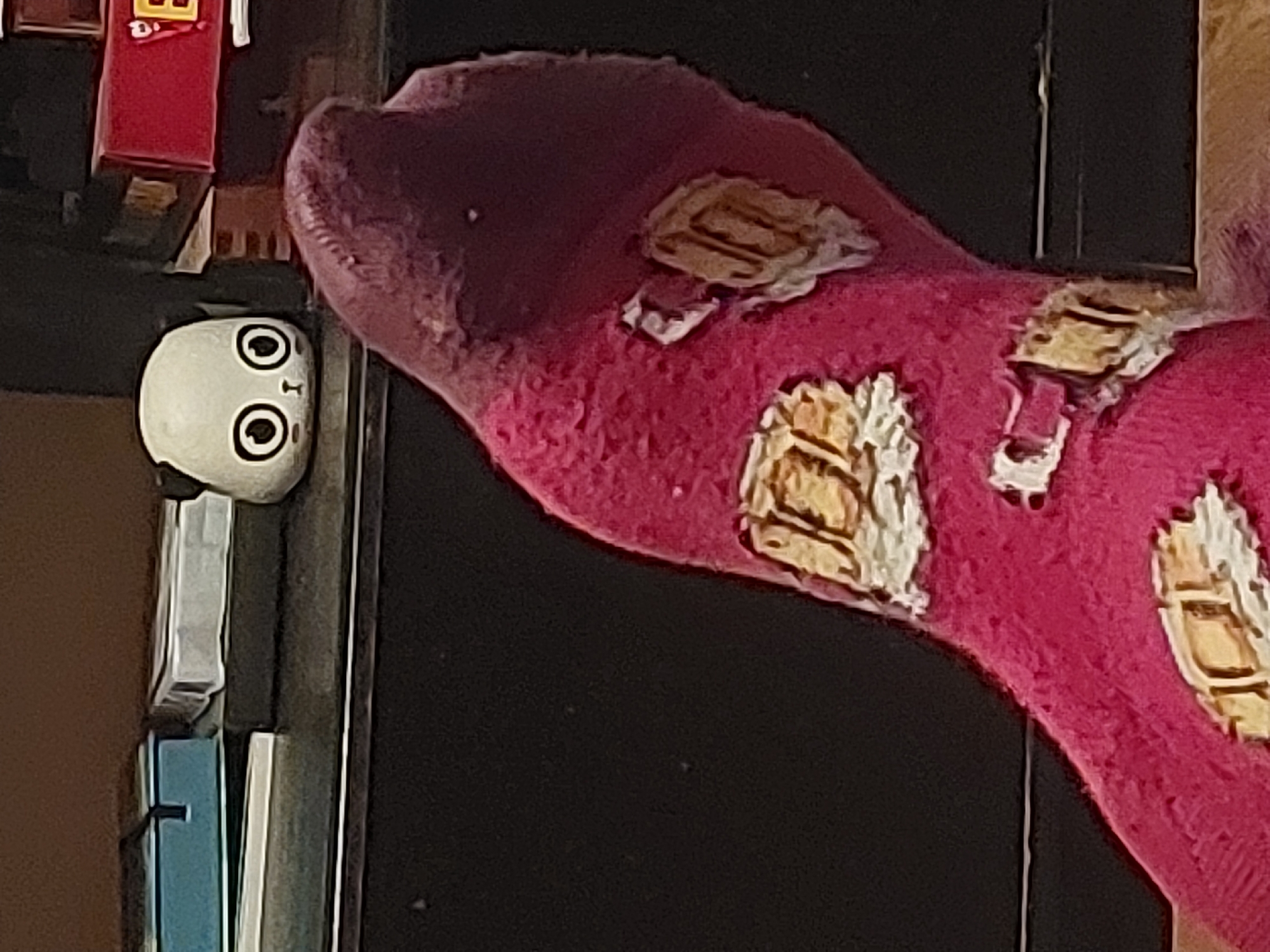 A close-up shows a white, cylindrical object with a cartoonish face (large eyes and a small mouth) near a stack of books and a red object, possibly a box. To the right, a plush, pink, sock-like object with yellow and white patches partially obscures a dark background, likely a wall. The scene appears indoors. The city and country are not evident in this image.