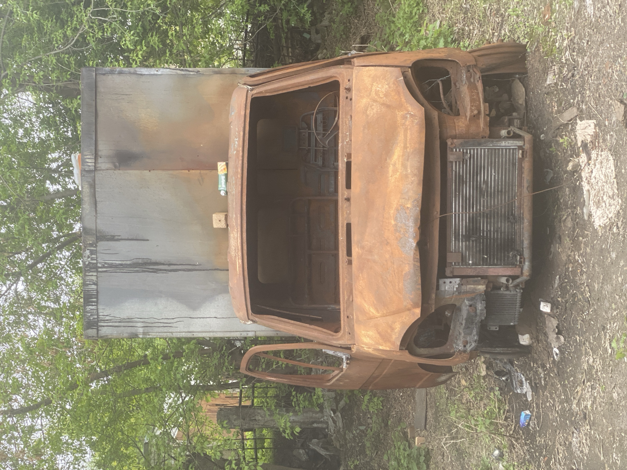 A burnt-out truck is situated in a grassy area, likely on the outskirts of a city. The vehicle is heavily rusted and lacks significant portions of its body, revealing the interior framework. The truck's radiator is visible at the front. The back of the truck appears to be a gray, box-shaped cargo area, heavily damaged by soot. Vegetation including trees and grass surround the vehicle. Based on prior knowledge, this is located in Novokuznetsk, Russia.