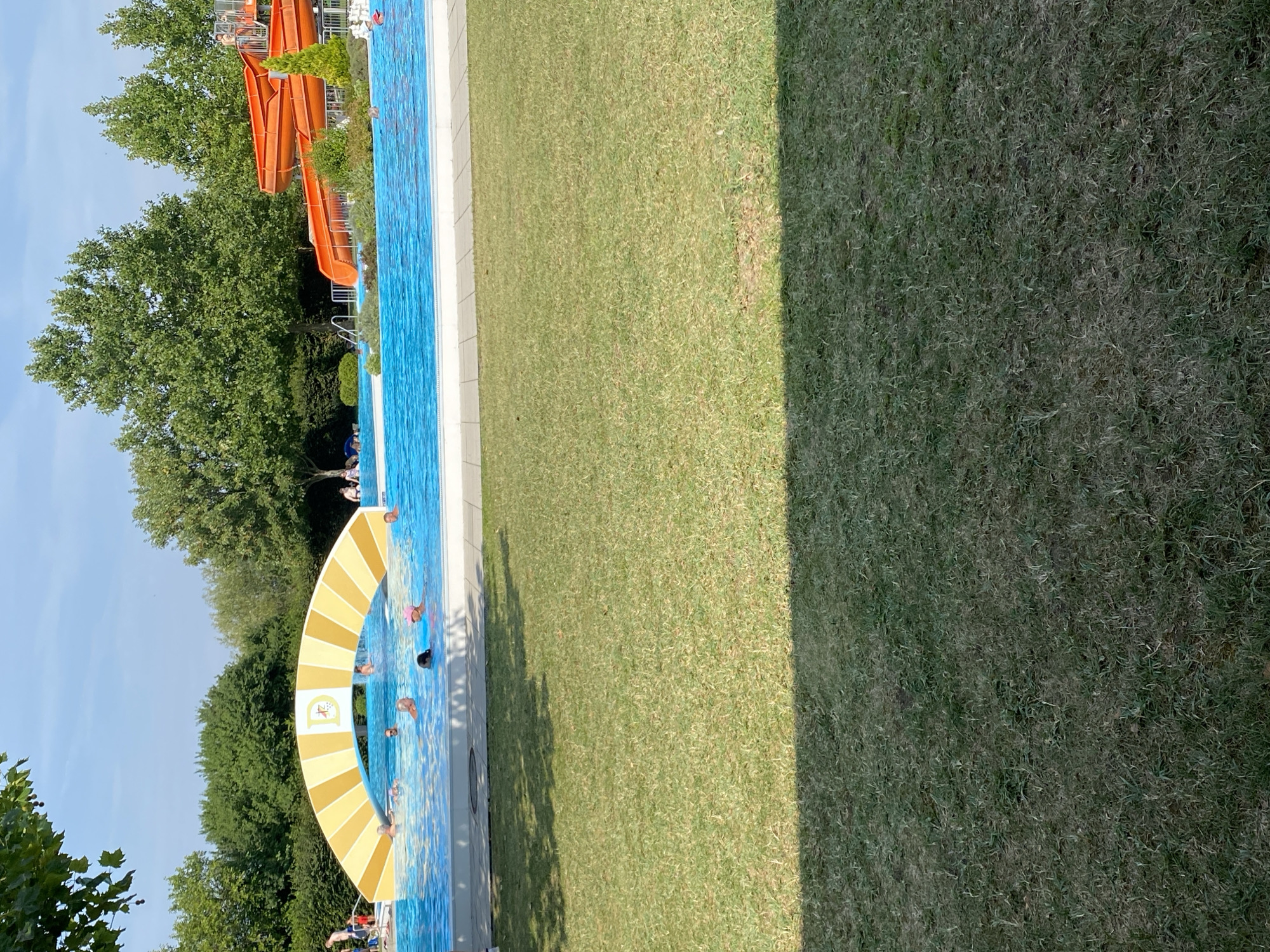 The image depicts an outdoor swimming pool area in Deutschkreutz, Austria. In the foreground, a well-maintained lawn is visible, with shadows cast by unseen trees. The middle ground features a bright blue swimming pool with several people swimming and wading. An arched structure, yellow and white striped with a central logo, spans part of the pool. In the background, vibrant green trees form a dense backdrop. To the right, a large orange waterslide complex is prominent. The sky above is clear and blue.