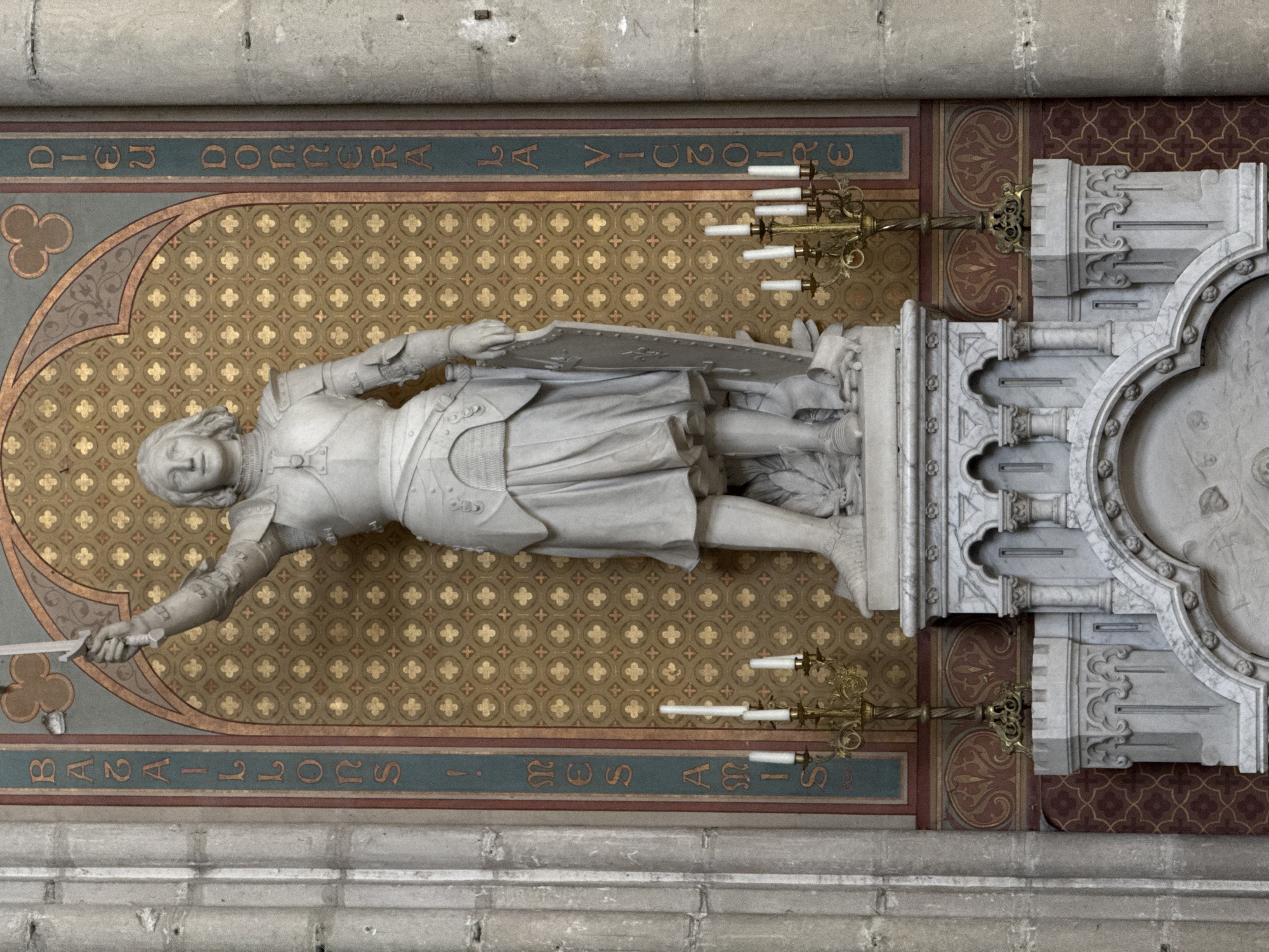 The image shows a marble statue of Joan of Arc, located in the Cathedral of Saint Peter and Saint Paul in Nantes, France. She is depicted as a young woman in armor, holding a sword aloft in her right hand and a shield in her left. The statue stands on a marble pedestal adorned with Gothic architectural elements. Behind the statue, a wall is decorated with a patterned tapestry and two vertical inscriptions in French. The inscription on the left reads "BATAILLONS MES AMIS", and the inscription on the right reads "DIEU DONNERA LA VICTOIRE". Two candelabra with lit candles are placed on either side of the pedestal. The statue is situated within the interior of the cathedral, with stone columns visible on the left and right sides of the frame. The overall lighting suggests an indoor setting, possibly illuminated by ambient light and the candles.