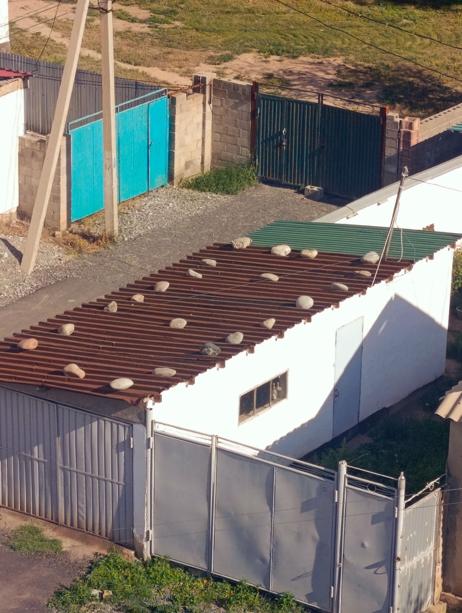 The image shows a view from above of a residential area in Naryn City, Kyrgyzstan. In the foreground, a long, low white building with a corrugated metal roof is visible. The roof has several large stones placed on it. The building has a single door and a small window with a barred opening. A grey metal fence runs along the front of the building. To the left of this building, a narrow driveway or path is visible, bordered by a chain-link fence and a brick wall. Further left, there are two turquoise gates set within a brick wall. Behind the white building, there is a green corrugated metal roof section, possibly an extension or a separate structure. A concrete utility pole stands to the left of the turquoise gates. The background consists of dry grass and more fencing. The scene appears to be a backyard or an alleyway between properties.