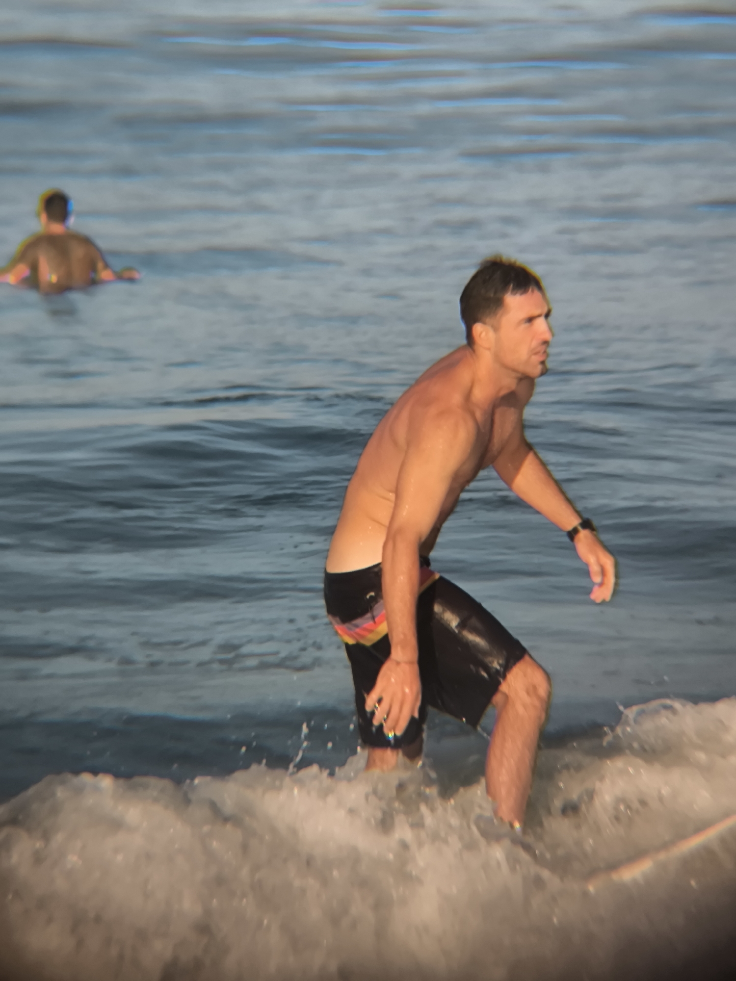 The image shows a man standing in shallow water with a wave washing around his legs. He is wearing dark swim trunks with a colorful waistband and a watch on his left wrist. His torso is bare. In the background, another person is seen waist-deep in the water. The location appears to be a beach, possibly in North Kuta, Indonesia, judging by the likely coastal setting and the people's attire.