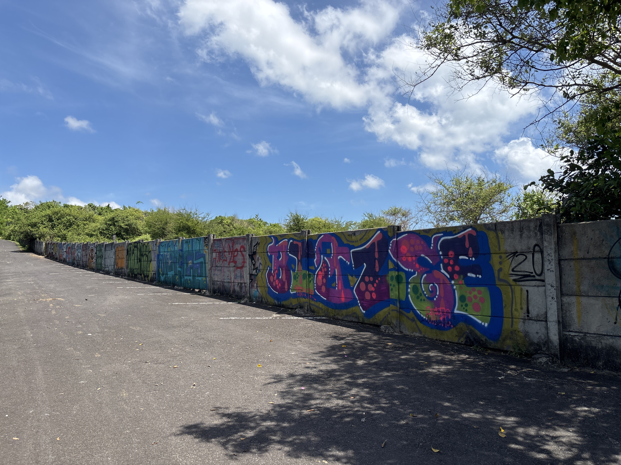 The image depicts an outdoor scene in an unknown location in Indonesia. An asphalt road stretches into the distance along a long, gray concrete wall. The wall is covered in graffiti, varying in color and style, from simple tags to more complex designs. The road appears to be a parking area, marked by faded white lines. Overhead, a blue sky with scattered white clouds is visible. Lush green vegetation lines the top of the wall, creating a backdrop to the scene. The shadows cast by trees fall onto the road surface. No people are present. The overall environment suggests an urban or semi-urban area with signs of both natural and human elements.