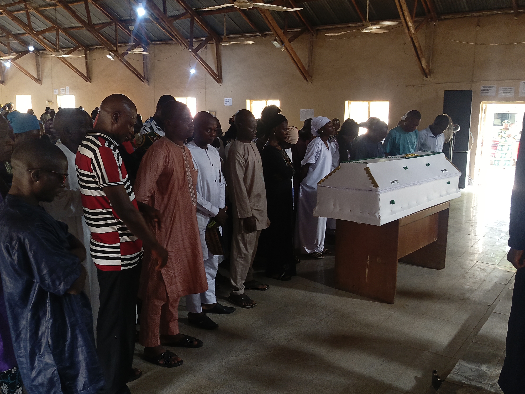Location: Interior of a building in Akwanga, Nigeria.
Scene: A funeral gathering.
People: A group of people stand around a casket. They are dressed in a variety of clothing, some wearing traditional Nigerian attire. Most are men, but there are some women present.
Objects: A white casket with green and gold accents rests on a wooden stand. The building has a high, exposed beam ceiling and multiple windows. Ceiling fans are visible.
Actions: The people are standing quietly, some with their heads bowed in respect or prayer.
Environment: The building appears to be a community hall or church. The room is well-lit and spacious.