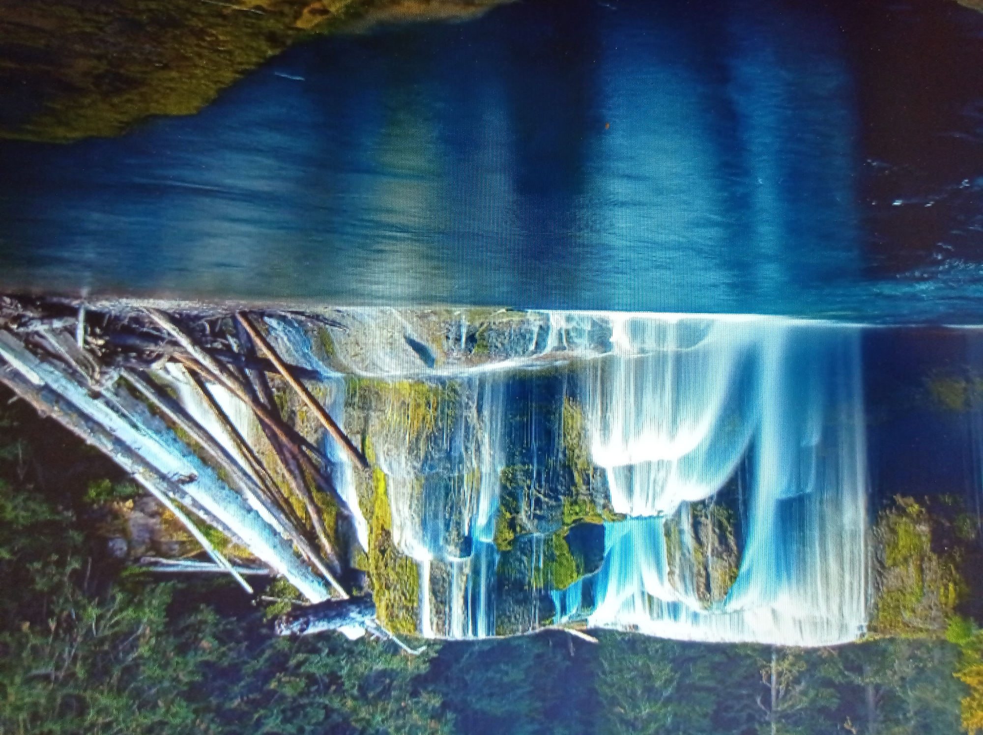 The image depicts a picturesque waterfall cascading into a calm, blue body of water. The waterfall is wide and segmented, with water flowing over moss-covered rocks and ledges. The surrounding environment is lush and green, with dense trees visible behind the waterfall and on its sides. A collection of fallen logs and branches is piled on the right side of the scene, near the edge of the waterfall. The water reflects the vibrant blues and greens of the surrounding landscape, creating a serene and natural setting. There are no people or animals visible in the image. The location is not in the city of Mukachevo, Ukraine.