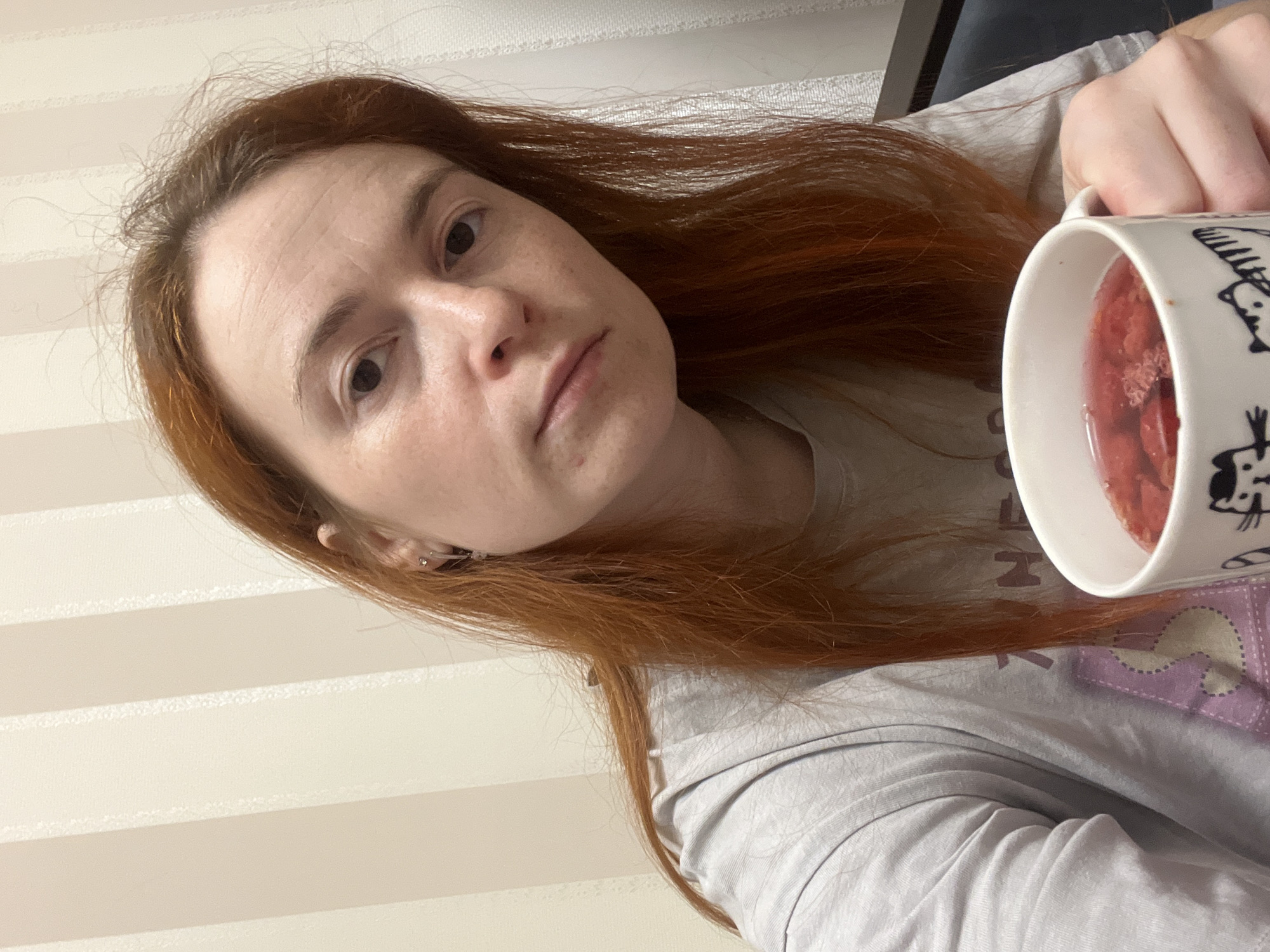 A woman with red hair is holding a white mug filled with a red liquid, possibly a fruit infusion or tea, towards the camera. She has fair skin and is wearing a gray t-shirt. The background consists of vertical striped wallpaper. The location, Chaikovsky, Russia, is not discernible from the image.