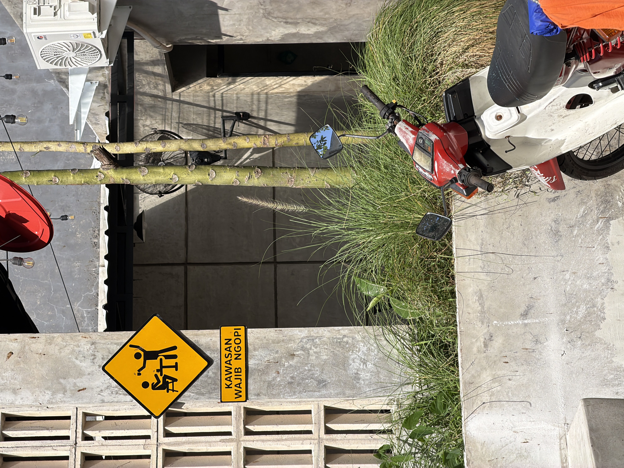 The image depicts an outdoor scene in Kuta Selatan, Indonesia. The foreground features a low concrete wall with decorative vents. A yellow diamond-shaped sign showing a stick figure pushing another stick figure in a chair is attached to the wall next to a sign reading "KAWASAN WAJIB NGOPI." Behind the wall, tall grasses and vegetation grow near a parked white moped with a red front section. A metal pipe with branching stalks runs above the wall, near an outdoor seating area with a red umbrella, string lights, and an air conditioning unit mounted on a nearby building. The background shows sections of concrete buildings and shadowed areas.