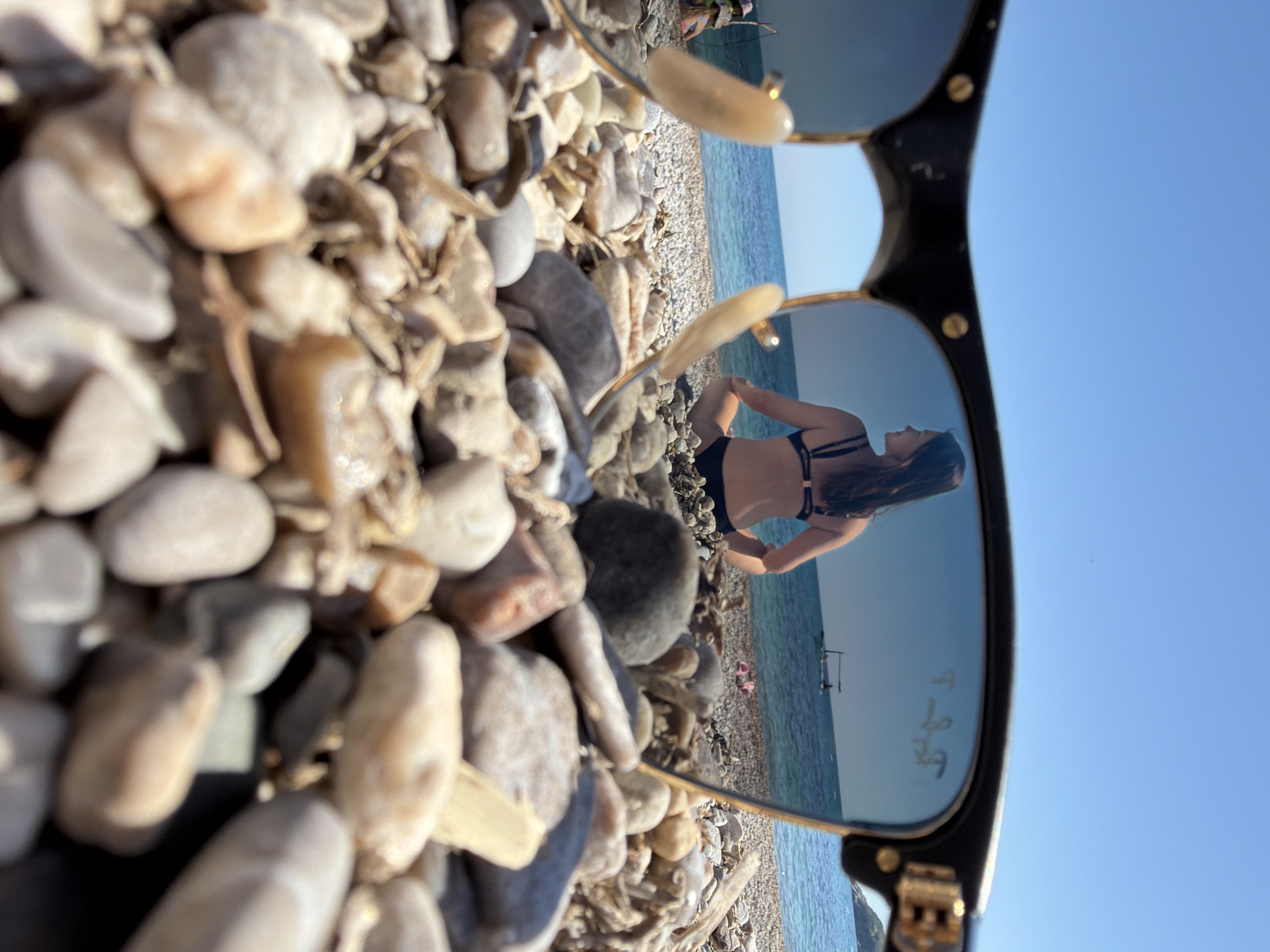 A close-up shot focuses on a collection of pebbles and sand, with the lens of a pair of sunglasses acting as a reflective surface, capturing a scene of a woman in a black bikini standing on a pebble beach in Prijevor, Montenegro. The ocean is visible behind her, merging with the blue sky, creating a seaside backdrop. The sunglasses themselves are the primary subject, with their reflection providing context to the surrounding environment.