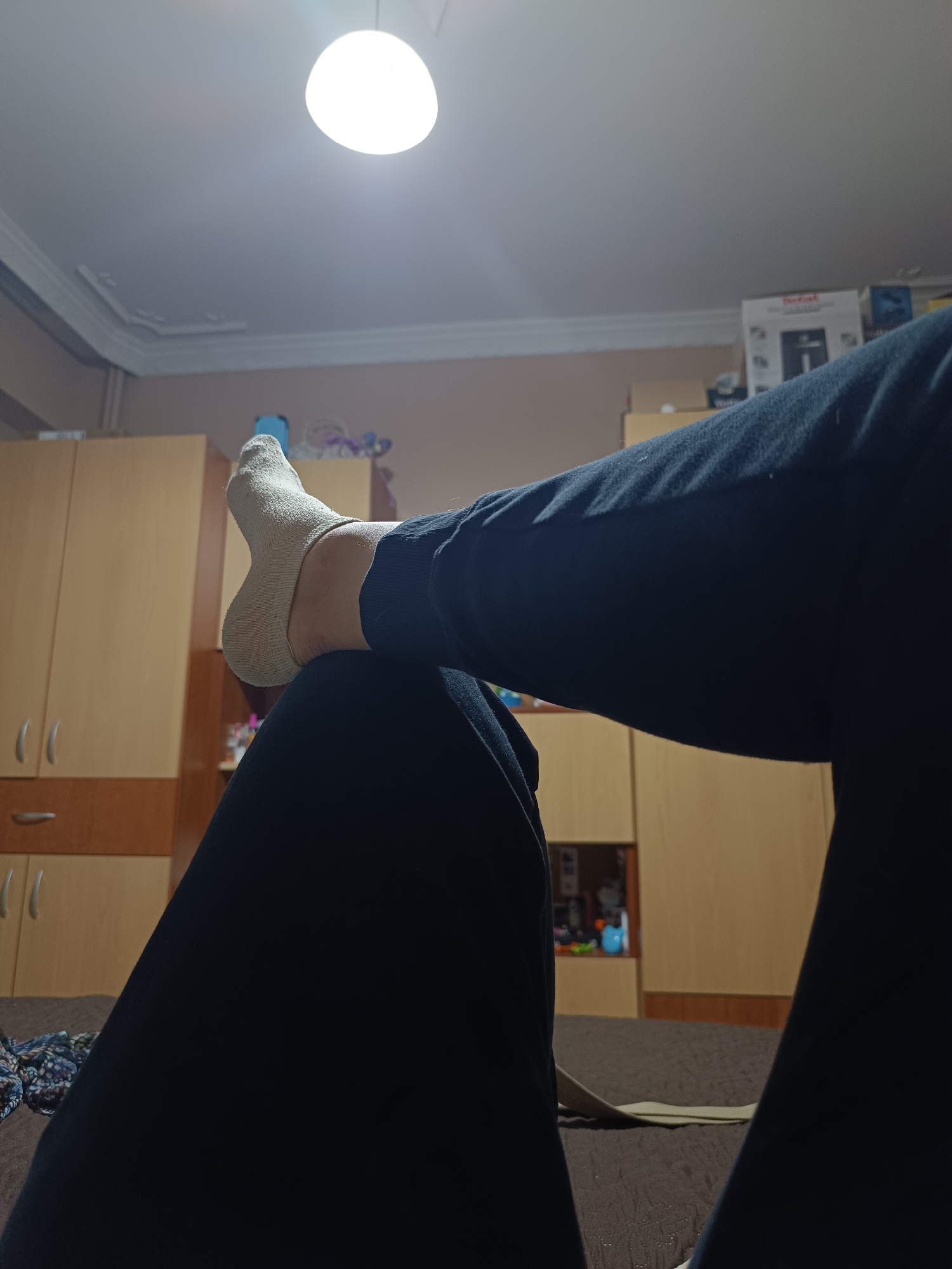 A low-angle, indoor shot focuses on a person reclining on a bed, with their legs raised. The person is wearing dark blue pants, and their left foot is visible, clad in a light-colored sock. The foot rests on the person's right leg.
Behind the legs, light brown wooden cabinets and shelves line the wall. A few items are visible on the shelves. In the upper right corner, a white box with the word "Tefal" can be seen. A ceiling light illuminates the room.
The location is Pleven, Bulgaria.