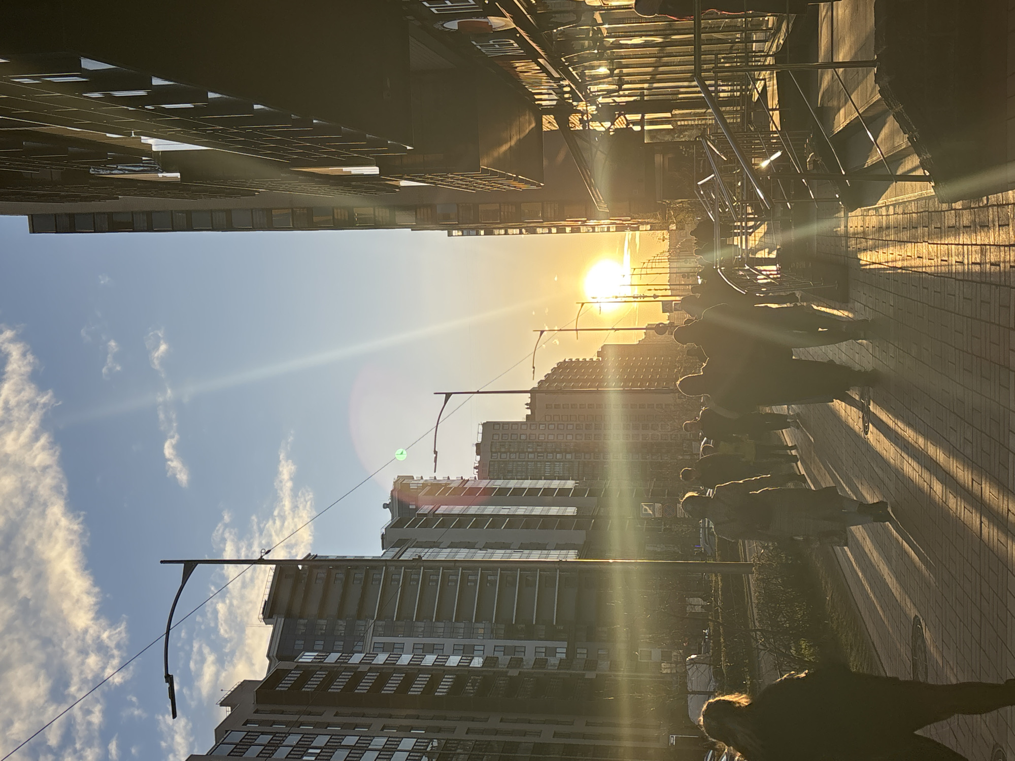 The image depicts an urban street scene in Murino, Russia, at sunset, filled with warm, golden light. Tall, modern apartment buildings line both sides of the street, showcasing numerous windows and balconies. The low setting sun, a bright golden orb, is prominently positioned towards the right side of the horizon, casting intense light rays and noticeable lens flares across the frame.
A wide, paved sidewalk runs along the right side, extending into the distance and bordered by a metal railing. Several individuals are seen walking along this path, their figures largely silhouetted against the bright backlight of the sun. They cast long, dramatic shadows that stretch across the sun-drenched pavement, emphasizing the late hour. A road is visible beyond the sidewalk, with some parked cars further down.
The sky on the left side of the image is a clear blue, dotted with scattered white clouds. Streetlights with distinctive curved poles are spaced along the thoroughfare. Patches of green vegetation, including trees and grass, are visible between the buildings and the street. The overall atmosphere is serene and warm, highlighted by the dramatic sunset lighting. While some indistinct text or signage is present on a building in the mid-ground, it is not clearly legible.