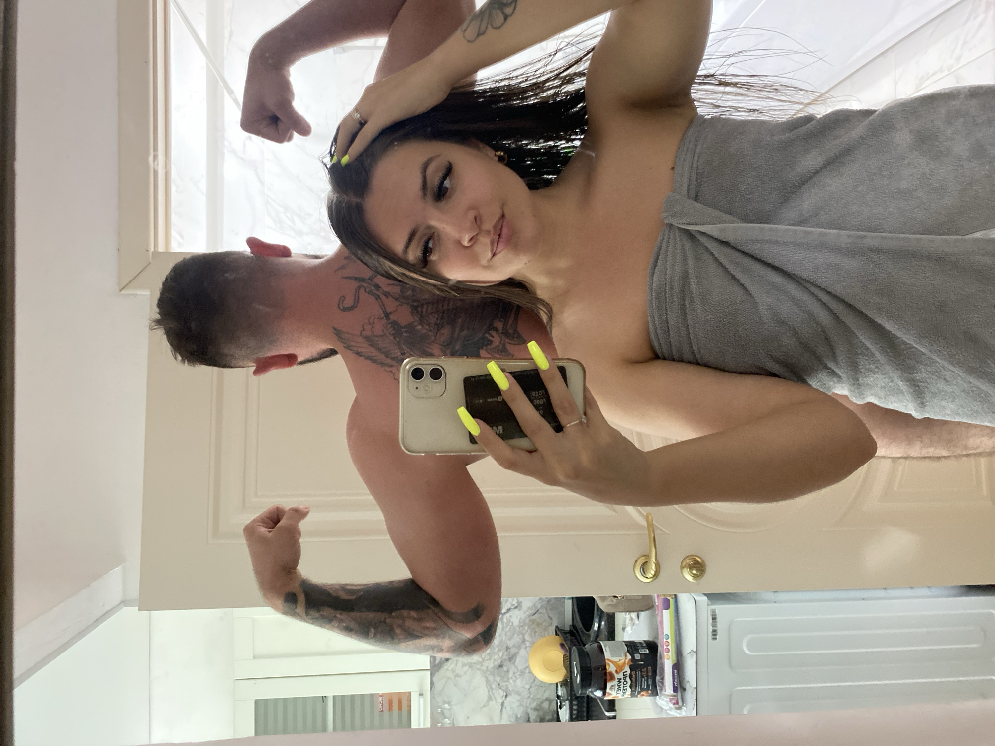 In a bathroom, a woman with long brown hair wrapped in a gray towel takes a selfie in a mirror. Her nails are painted neon yellow. Behind her, a man with tattoos on his arms and back flexes his bicep. In the reflection, a washing machine, a shelf with a protein supplement container, and a yellow spherical object are visible.