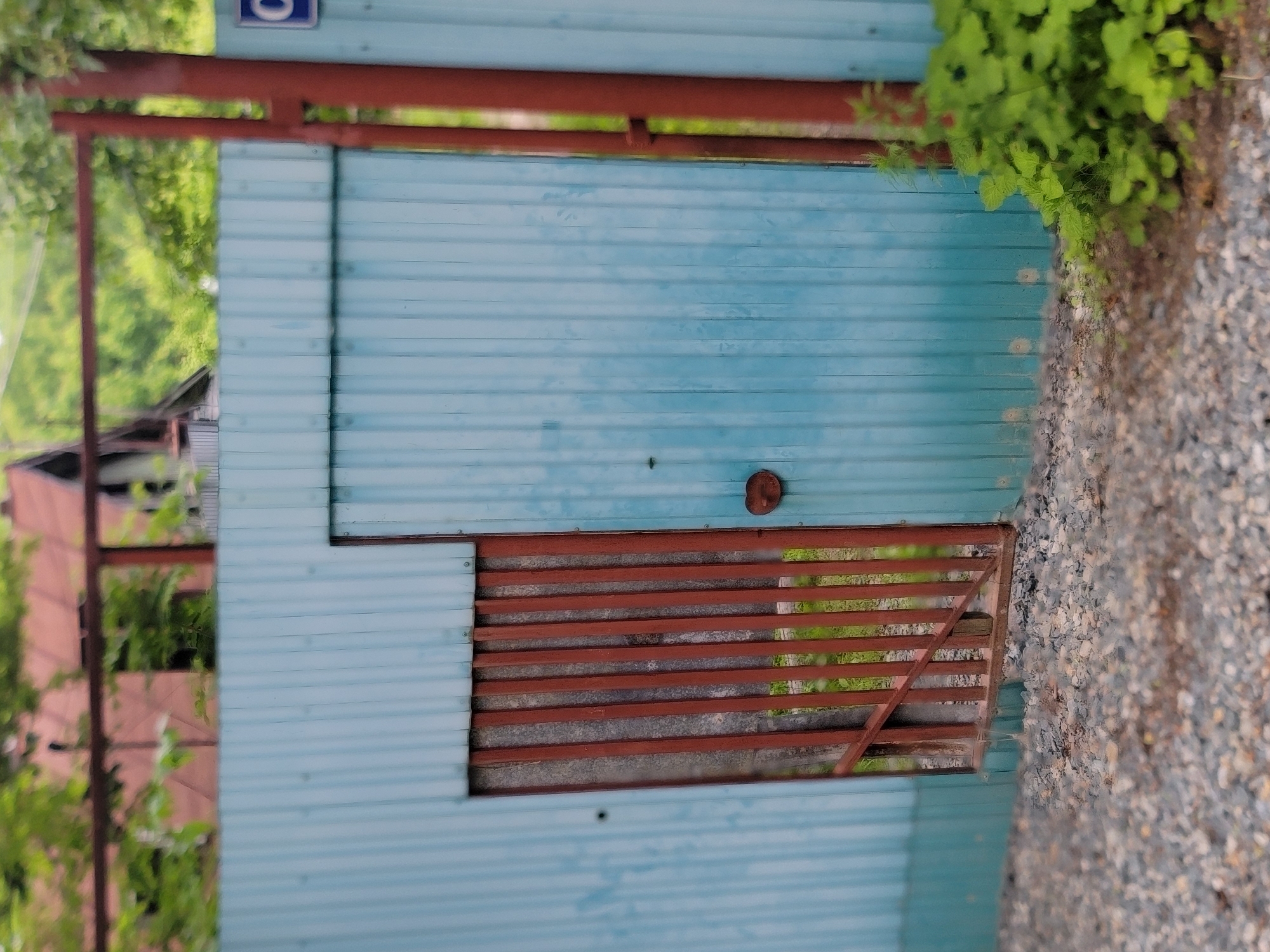 This is a close-up, eye-level, vertical, outdoor shot of a light blue corrugated metal fence. A small, rusted metal gate, approximately waist-high, is set within the fence on the left. The gate consists of vertical metal bars. The rest of the fence is solid. To the right of the gate, the fence forms a solid panel with a round, rusted metal handle near the bottom. A dark red, vertical metal pole is attached to the right edge of the fence, continuing upwards and backwards out of frame. In the background, out of focus, are green foliage, a reddish-brown sloped roof of a building, and possibly a portion of another structure. The ground in the foreground is covered with small grey gravel. On the right side of the image, some green foliage is growing near the base of the fence. A blue square sign with a white letter "C" is visible on the fence to the right of the vertical pole. The scene appears to be an exterior residential or industrial area in Vladivostok, Russia.