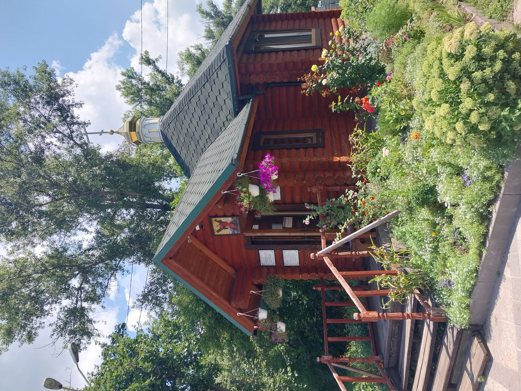 The image depicts a small wooden chapel with a golden dome and cross, situated in a garden setting in Dnipro, Ukraine. The chapel is constructed from logs and has a dark shingled roof. A wooden staircase with a railing leads up to the entrance. The garden is filled with a variety of flowers and greenery. In the background, trees and a blue sky with clouds are visible. A streetlamp is present to the left of the chapel. There are no people visible in the image.