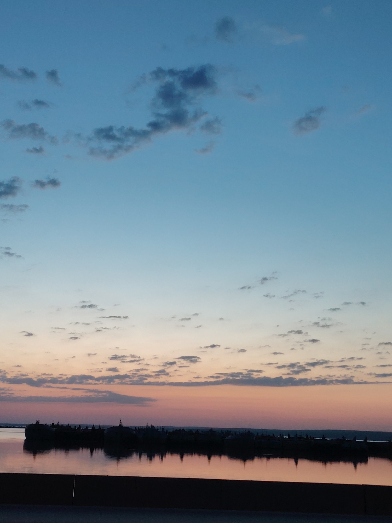 The image depicts a twilight scene in Chaikovsky, Russia. The sky exhibits a gradient of blues and pinks, transitioning from a darker blue at the top to a rosy hue near the horizon. Scattered clouds are visible throughout the sky. Below the horizon, a body of water reflects the colors of the sky. A dark, silhouetted landmass stretches across the middle ground, likely an island or the opposite shore. The outlines of structures, potentially buildings and trees, are visible on the landmass. A dark, horizontal shape occupies the very bottom of the image, likely a road or bridge.