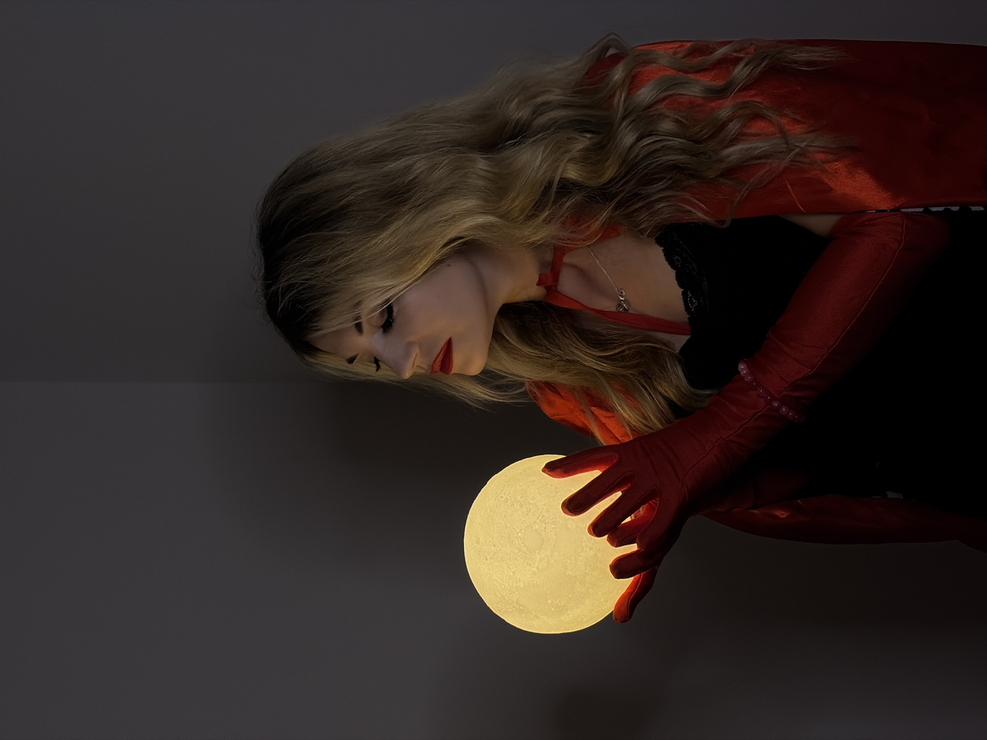 A blonde woman in red gloves and a red cape looks down at a glowing orb she is holding. She is wearing a black top and a red ribbon necklace with a small pendant. The background is a dark gray wall. The scene is indoors. There is no indication of Budva, Montenegro.