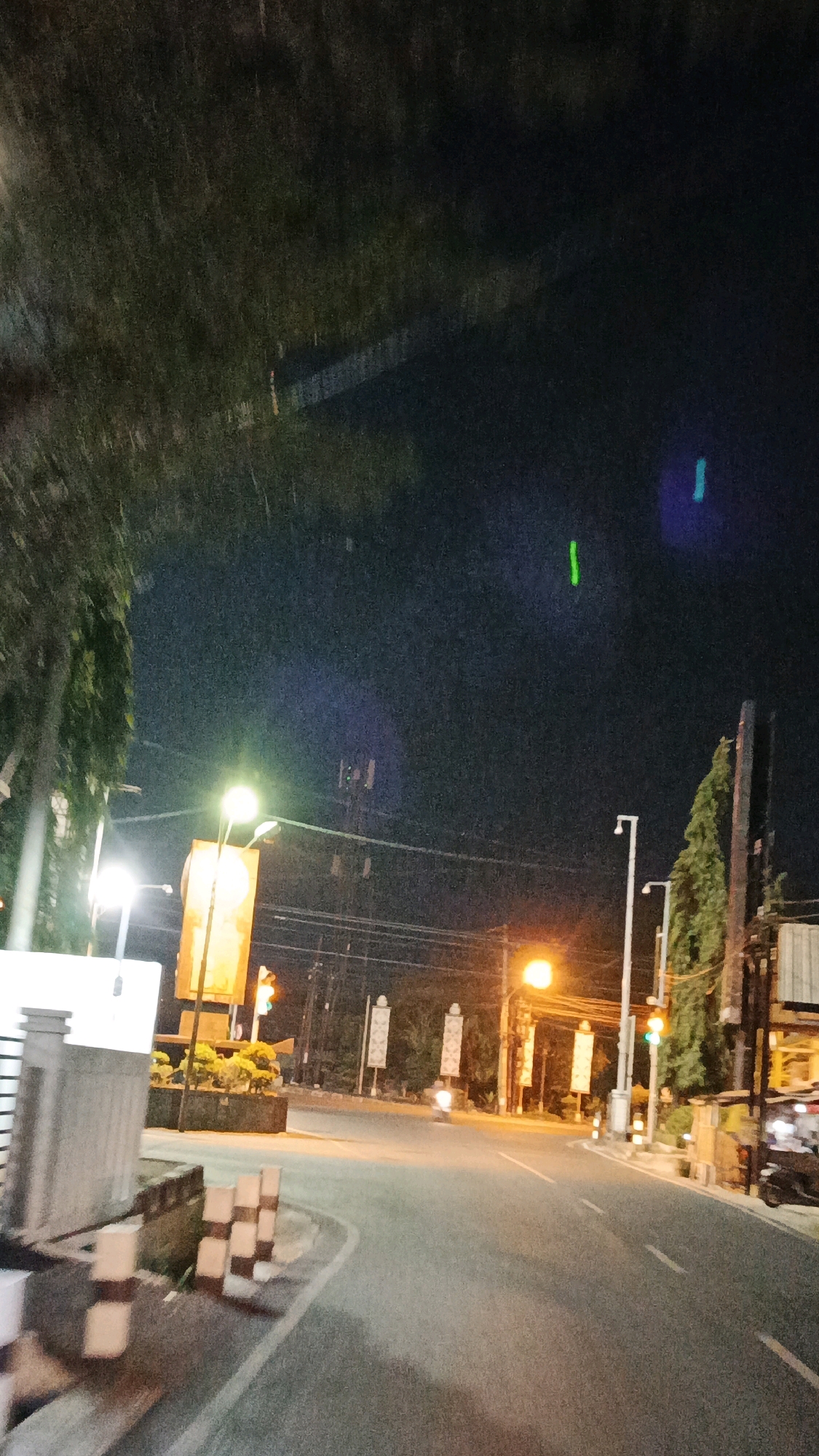 The image depicts a street scene at night in Surakarta, Indonesia, with streetlights and traffic signals illuminating the road. The perspective suggests the photograph was taken from a moving vehicle, as indicated by the slight blur and the curvature of the road.
In the foreground, a portion of a grey concrete barrier with alternating white and brown vertical stripes is visible on the left side of the road. The road itself is paved and appears to be a main thoroughfare, with lane markings faintly visible.
Further down the road, a large, brightly lit orange sign is positioned between two streetlights. Traffic signals, showing green and red lights, are also visible. On the right side of the road, there is a line of trees, some of which are tall and slender, and a building with a light-colored facade. Several vertical banners or flags with geometric patterns are also erected along the side of the road.
The sky is dark, characteristic of nighttime, with some faint light pollution. There are also some light streaks or lens flares visible in the sky, possibly due to the camera's exposure to bright lights. The overall atmosphere is one of a busy urban environment at night.