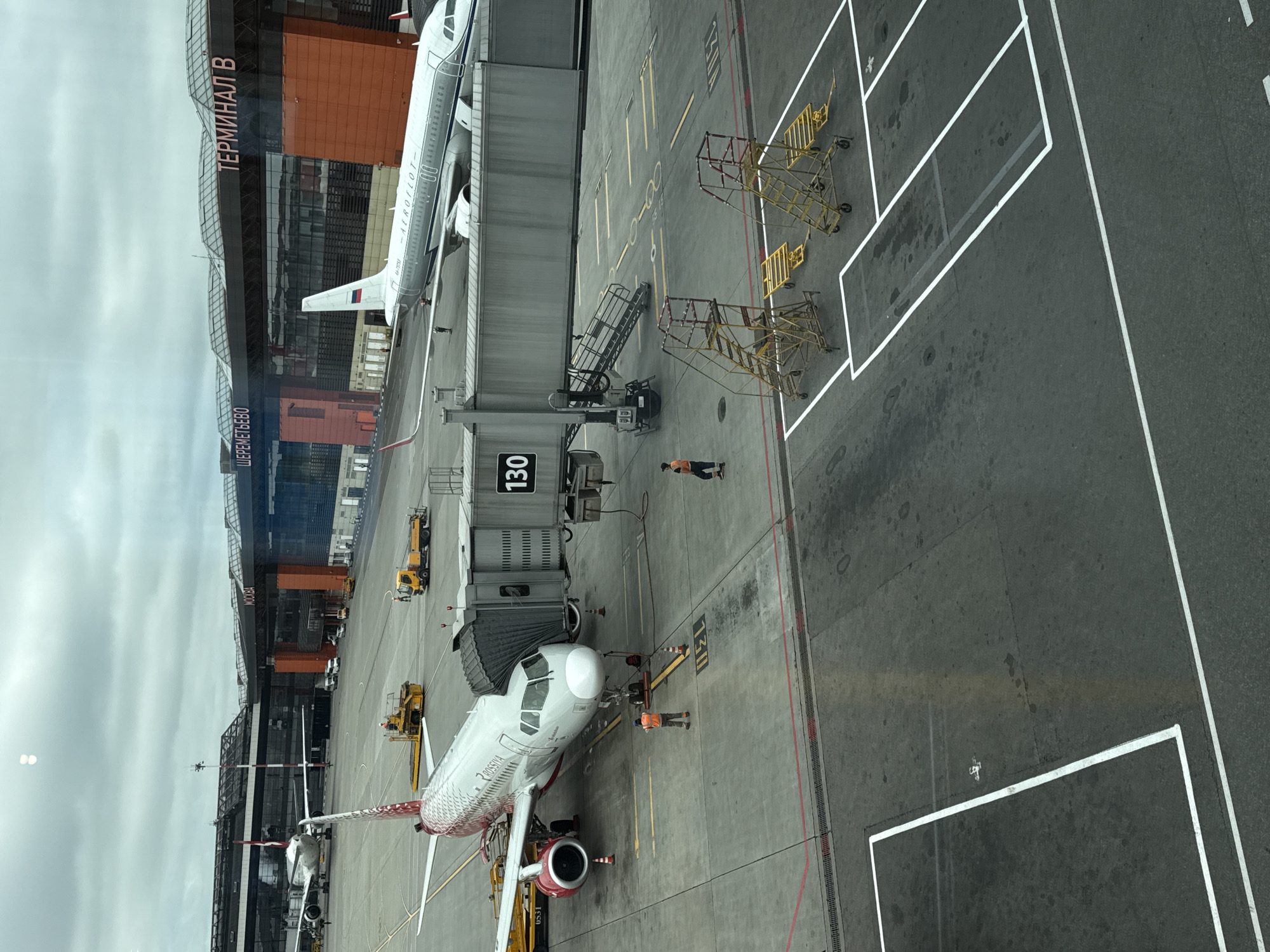 This is an aerial view of an airport tarmac at Sheremetyevo International Airport (SVO) in Rzhavki 2, Russia, specifically Terminal B. Arossia Airlines aircraft is parked at gate 130, connected to the terminal by a jet bridge. Two airport workers in high-visibility vests are on the tarmac near the aircraft. Another Aeroflot aircraft is parked in the background. Several service vehicles, including a tow tractor and a baggage cart, are visible on the tarmac. The sky is overcast.