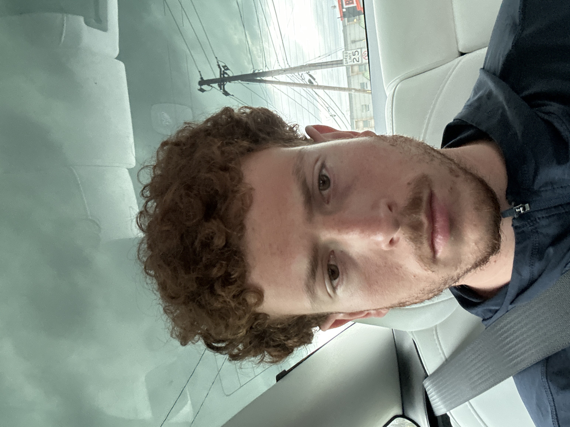 A light-skinned male with curly auburn hair and a beard is pictured from the chest up. He is wearing a dark-colored shirt and a gray seatbelt is visible across his chest. The background visible through the car window includes power lines, a building, and a street sign indicating a speed limit of 25 mph. The location is most likely in a residential or commercial area in the city of Orange, United States of America. The interior of the car has white seats and the window appears to be reflecting a cloudy sky.