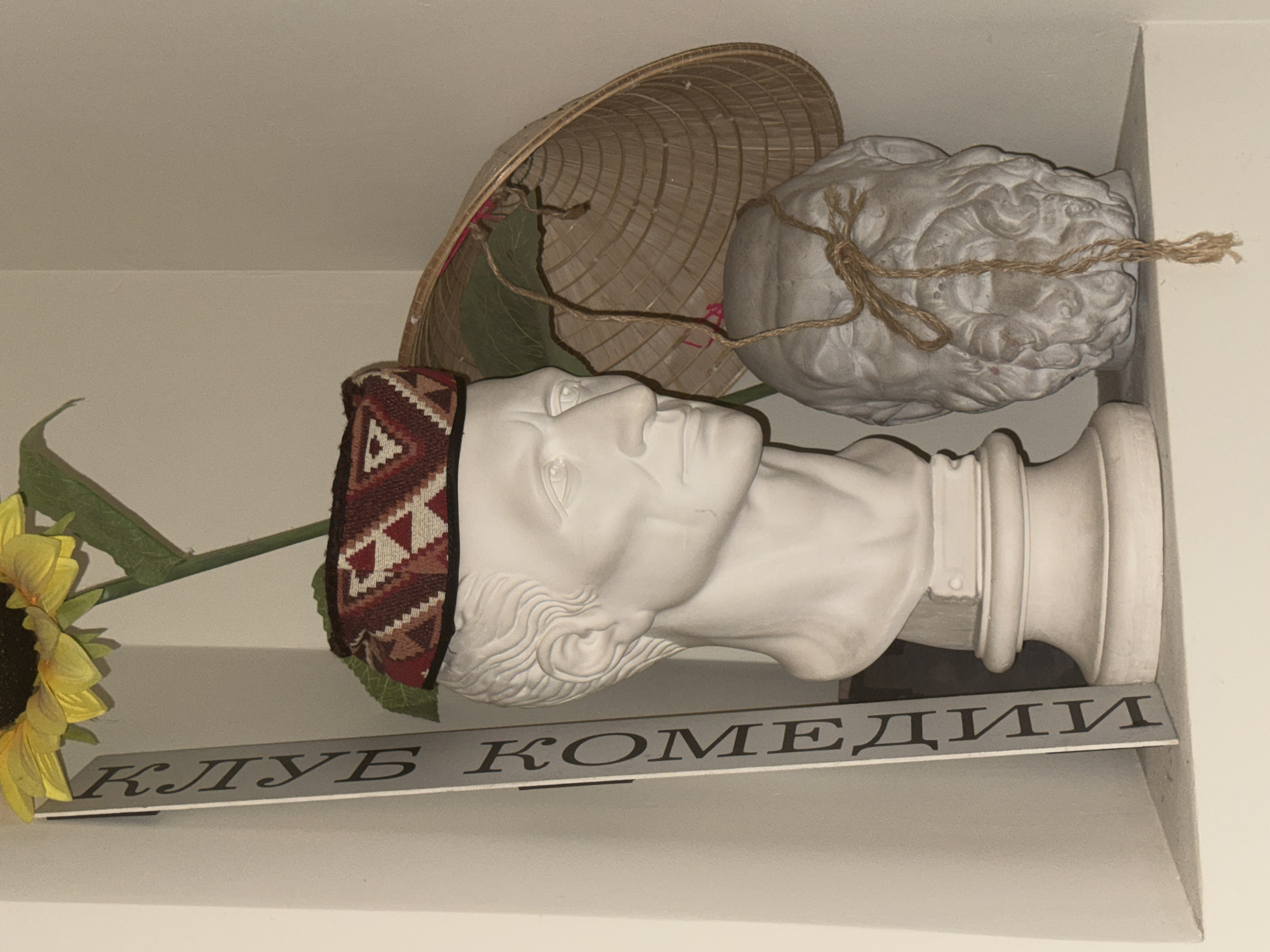 A still-life arrangement rests on a shelf in a room. A white bust of a man with a patterned headdress dominates the scene. To the left of the bust, a sunflower with green leaves leans towards the viewer. Underneath the bust, a sign bearing the Cyrillic inscription "КЛУБ КОМЕДИИ" (Comedy Club) is visible. Behind the bust, a conical Asian hat and a stone or concrete sculpture of a lion's head, bound with string, are positioned.
While the sign reads "Comedy Club," there are numerous comedy clubs in Moscow, Russia. The specific location of this image within Moscow is not identifiable from the provided visual information.