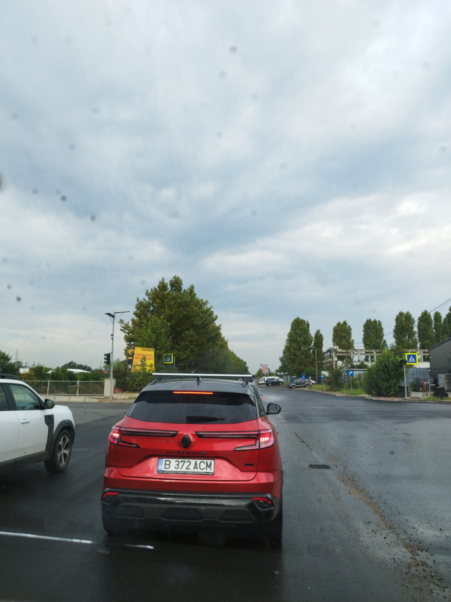 The image shows a view from inside a vehicle looking out at a city street in Bucharest, Romania. A red Renault Austral E-Tech Full Hybrid car with license plate B 372 ACM is prominently featured in the foreground. To the left, a white vehicle is partially visible. The street appears wet, suggesting recent rain. Traffic is present, with other vehicles in the distance. On the right side of the road, there are trees, buildings, and street signs, including a pedestrian crossing sign. The sky is overcast with clouds. The overall scene depicts a typical urban street environment during daylight hours, possibly in the afternoon.
