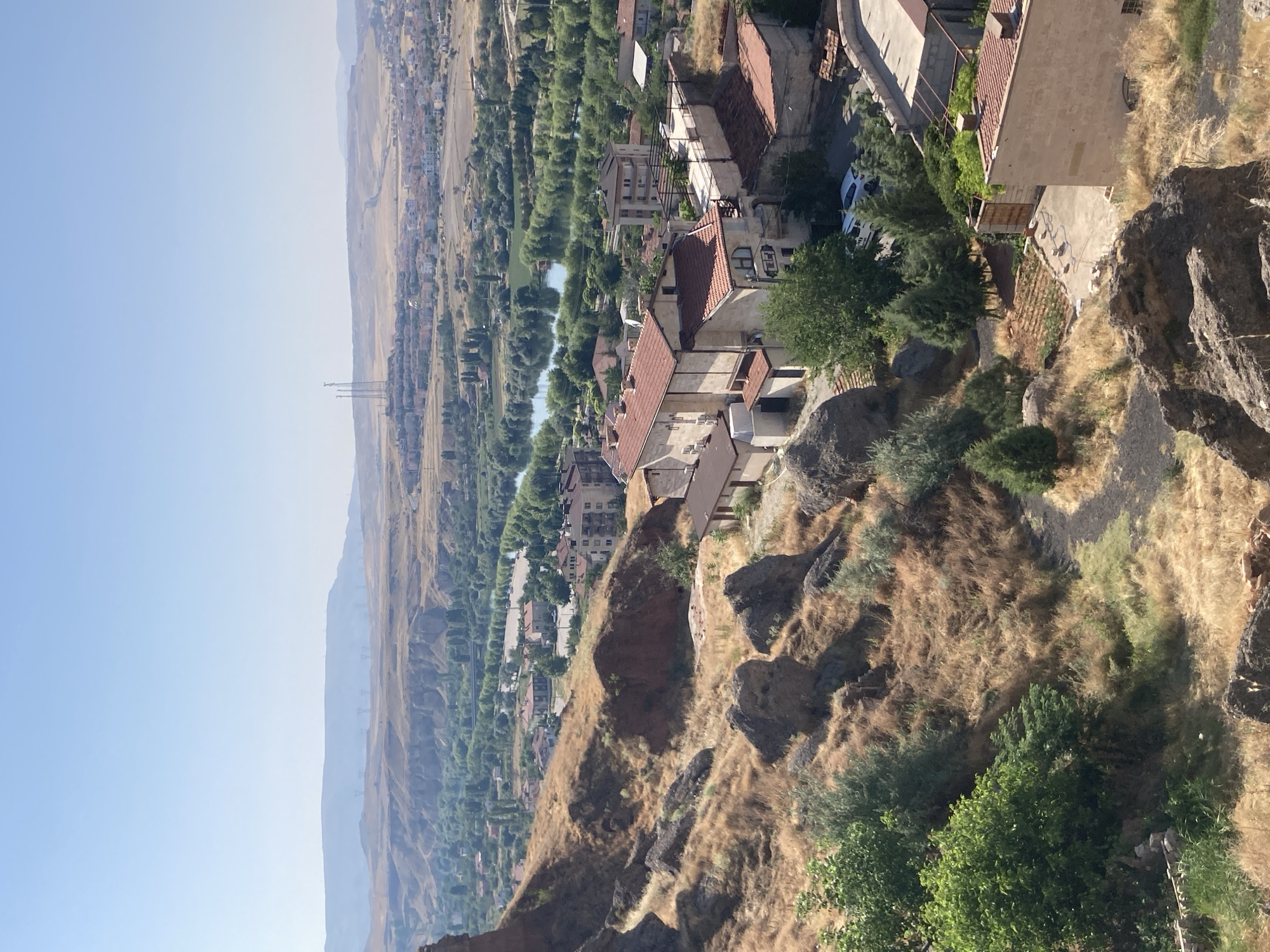 This is a high-angle, full-shot, vertical, outdoor, daytime, sunny photograph of the Avanos İlçe Merkezi, Turkey. The foreground features a rugged, dry, grassy hillside with large rocks and sparse vegetation. In the mid-ground, a town is visible with buildings featuring red-tiled roofs and cream-colored walls. A river winds through the landscape, lined with trees. In the distance, rolling hills and mountains form the horizon under a clear blue sky. A cluster of tall, lattice-like towers stands on a hill in the far distance, with wind turbines also visible on the distant hills.