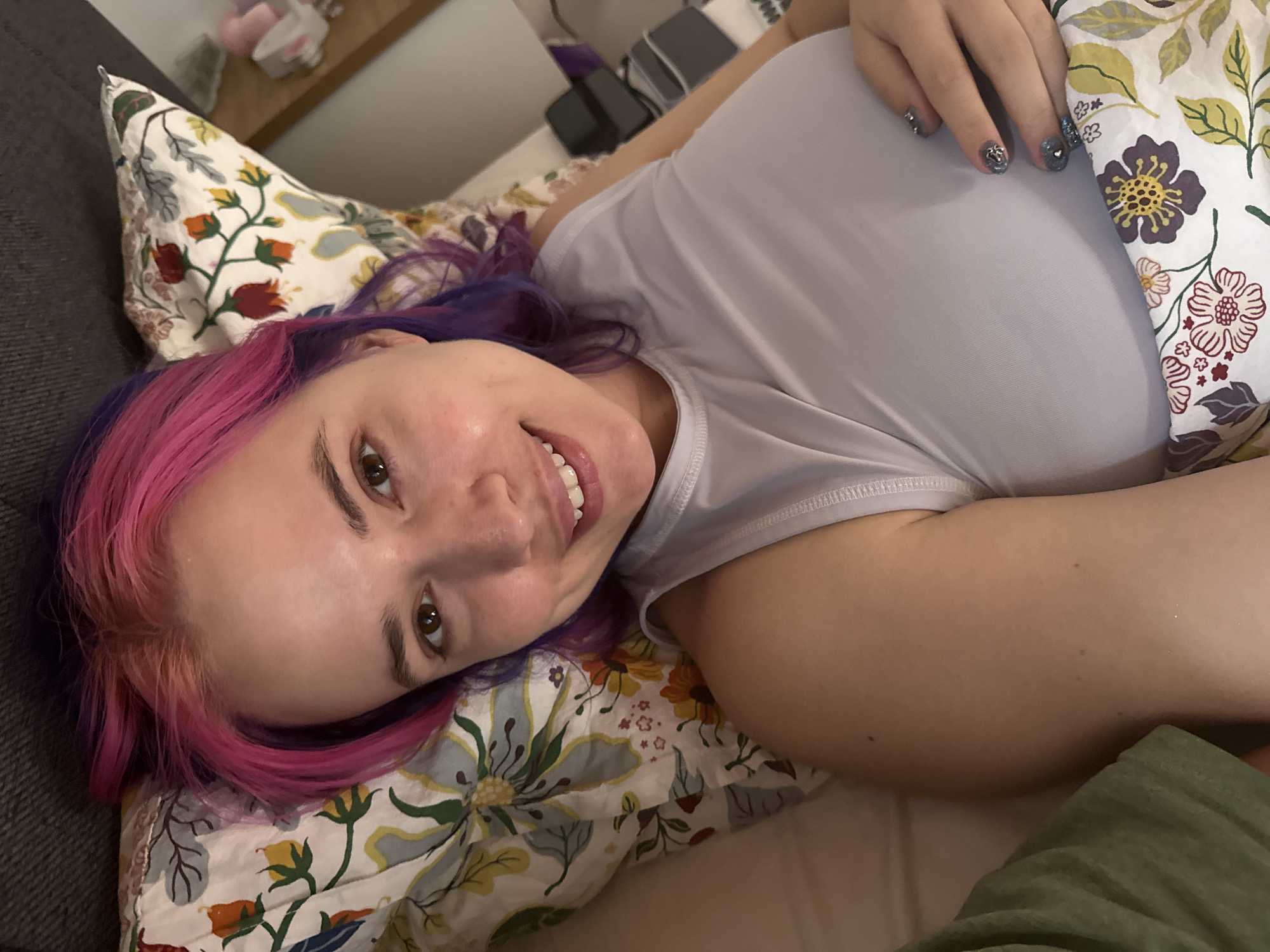 A woman with pink and purple hair is lying on a bed with floral patterned pillows and sheets. She is wearing a light gray sleeveless top and is smiling at the camera. Her left hand is resting near her head, showing off nails painted with a dark color and decorative embellishments. The location cannot be determined definitively but is potentially in City of Zagreb, Croatia. The environment is indoors, likely a bedroom.