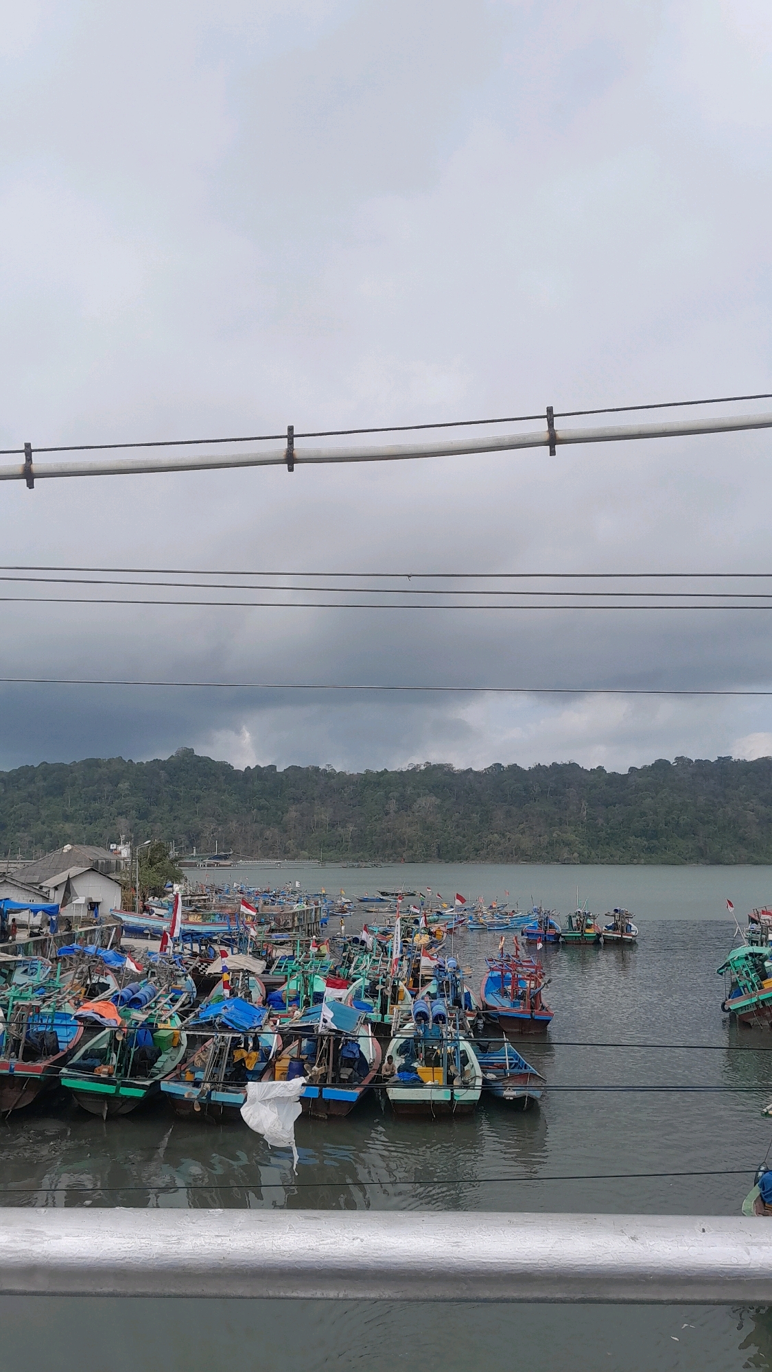 A medium-shot, vertical, eye-level, outdoor, handheld, medium-angle photograph captures a busy harbor filled with numerous small fishing boats docked in the waters of Cilacap, Indonesia. The sky is overcast and grey, with low-hanging clouds dominating the upper portion of the image. In the background, a densely forested hillside rises from the shoreline, with a few buildings visible amongst the trees. The foreground is occupied by a metal railing, suggesting the photo was taken from a bridge or elevated walkway.
The harbor is densely packed with dozens of small, colorful fishing boats, primarily painted in shades of green, blue, and red. Many of the boats are adorned with Indonesian flags, indicating national pride or perhaps a festive occasion. Some boats have tarps or covers, suggesting they are either being repaired, loaded, or are simply not in use. There are at least five visible figures, small and indistinct, on or near some of the boats, likely fishermen or dockworkers. One figure appears to be sitting on the edge of a boat, while others are engaged in activities that are not clearly discernible from this distance. A crumpled white piece of material, possibly plastic or fabric, is floating on the water in the middle ground, near the boats. The water appears calm, with subtle ripples indicating a gentle current. The overall scene depicts a working fishing port under a cloudy sky.