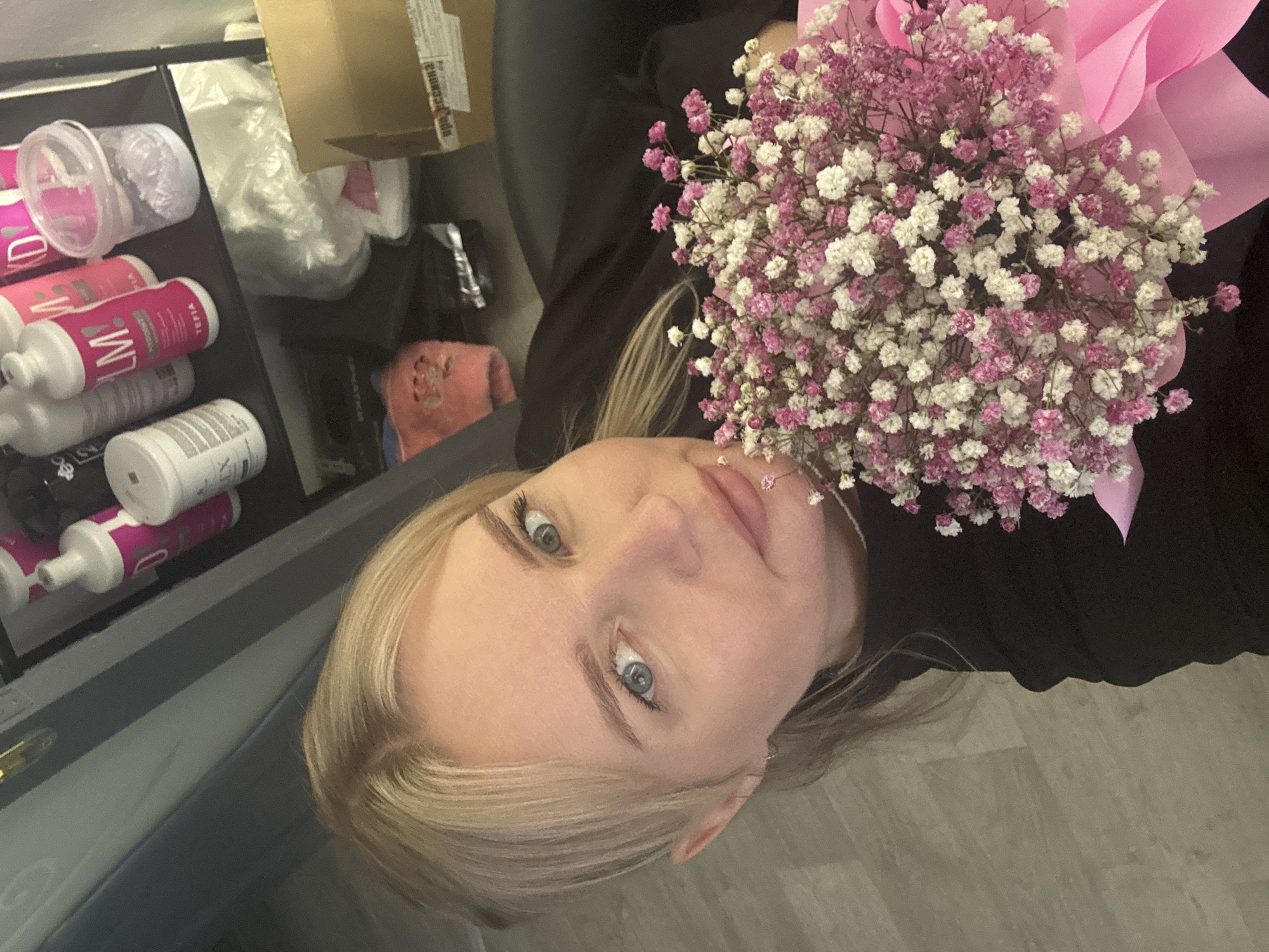 A woman with blonde hair and blue eyes holds a bouquet of pink and white baby's breath flowers wrapped in pink paper. She is wearing a black top. To the left of her is a shelving unit containing hair care products, including white bottles with pink labels and transparent containers with pink liquid. There is also a cardboard box and a pink towel. The location is identified as Chaikovsky, Russia.