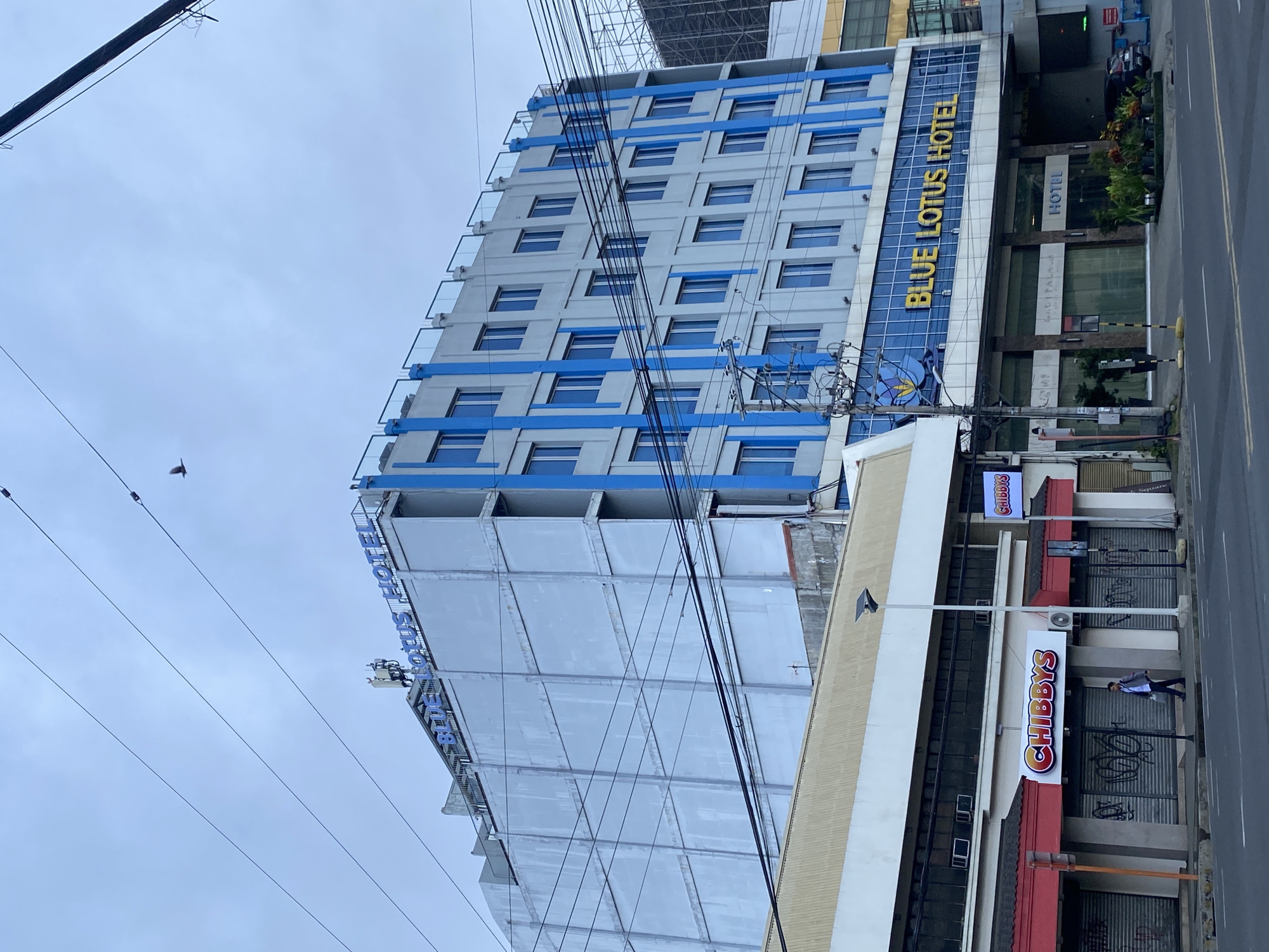 The image shows a wide shot of a street in Davao City, Philippines, featuring the Blue Lotus Hotel and several ground-level establishments. The Blue Lotus Hotel is a multi-story building with a light-colored facade and striking blue vertical accents. The hotel's name is prominently displayed in yellow lettering on the front. Below the hotel, a row of shops is visible. One of these shops is clearly labeled "Chibbys" with a bright, illuminated sign. The windows of the Chibbys store are covered with roller shutters, some of which have graffiti. A single person, dressed in a light jacket and dark pants, is walking on the sidewalk in front of the Chibbys establishment. Overhead, a complex network of electrical wires crisscrosses the frame. The sky is overcast. To the right of the Blue Lotus Hotel, another building with scaffolding is partially visible.