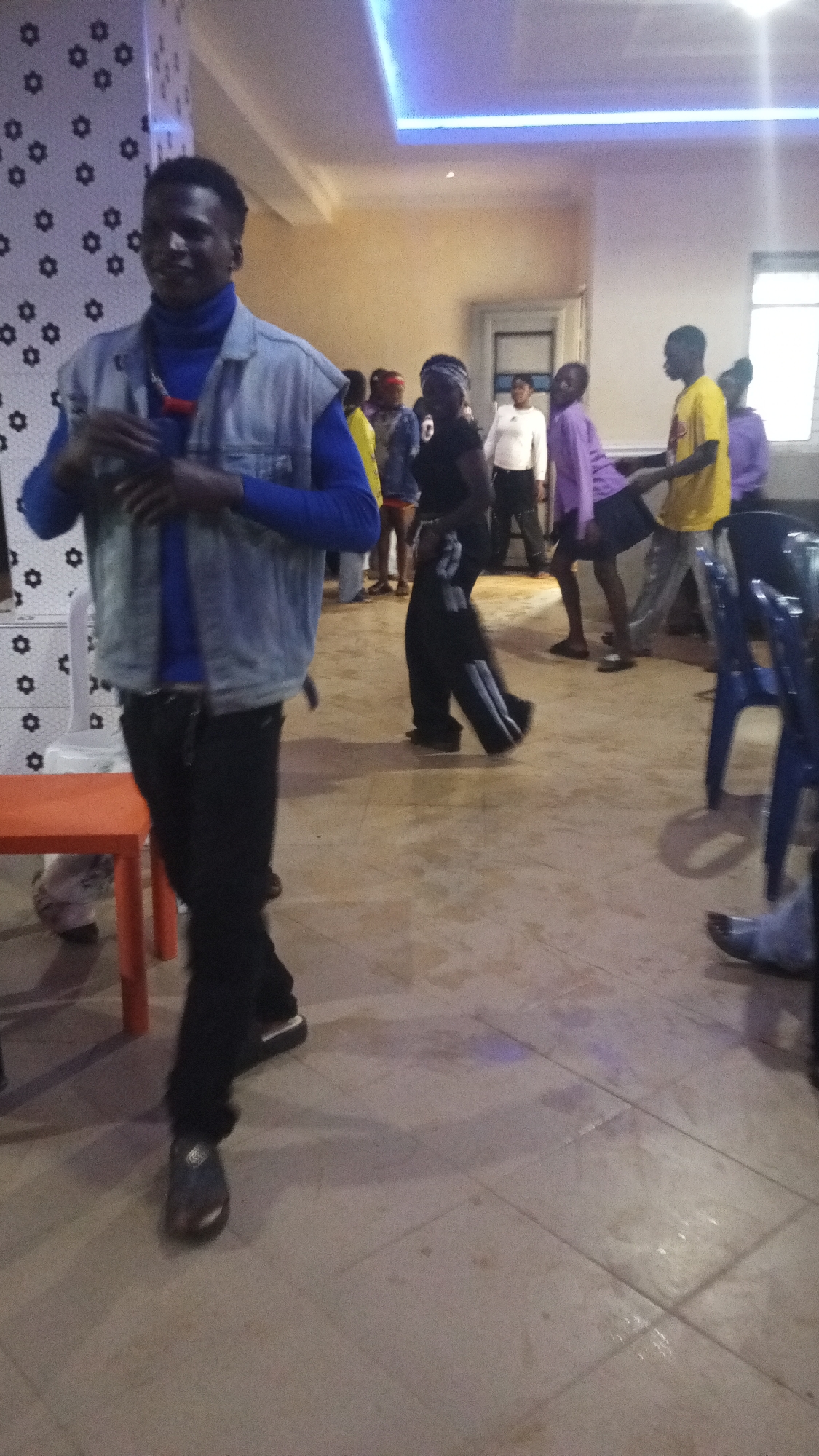 The image depicts an indoor scene with several individuals present. In the foreground, a person wearing a blue turtleneck and a denim vest is walking towards the camera. They are wearing black pants and dark shoes. To their right and further in the background, other people are visible, some appearing to be dancing or socializing. One individual in the background is wearing a yellow t-shirt and black pants, while another is in a purple long-sleeved shirt. The environment appears to be a room with tiled flooring and a ceiling with decorative lighting. There are chairs and a small orange table visible. The overall impression is that of a gathering or event.