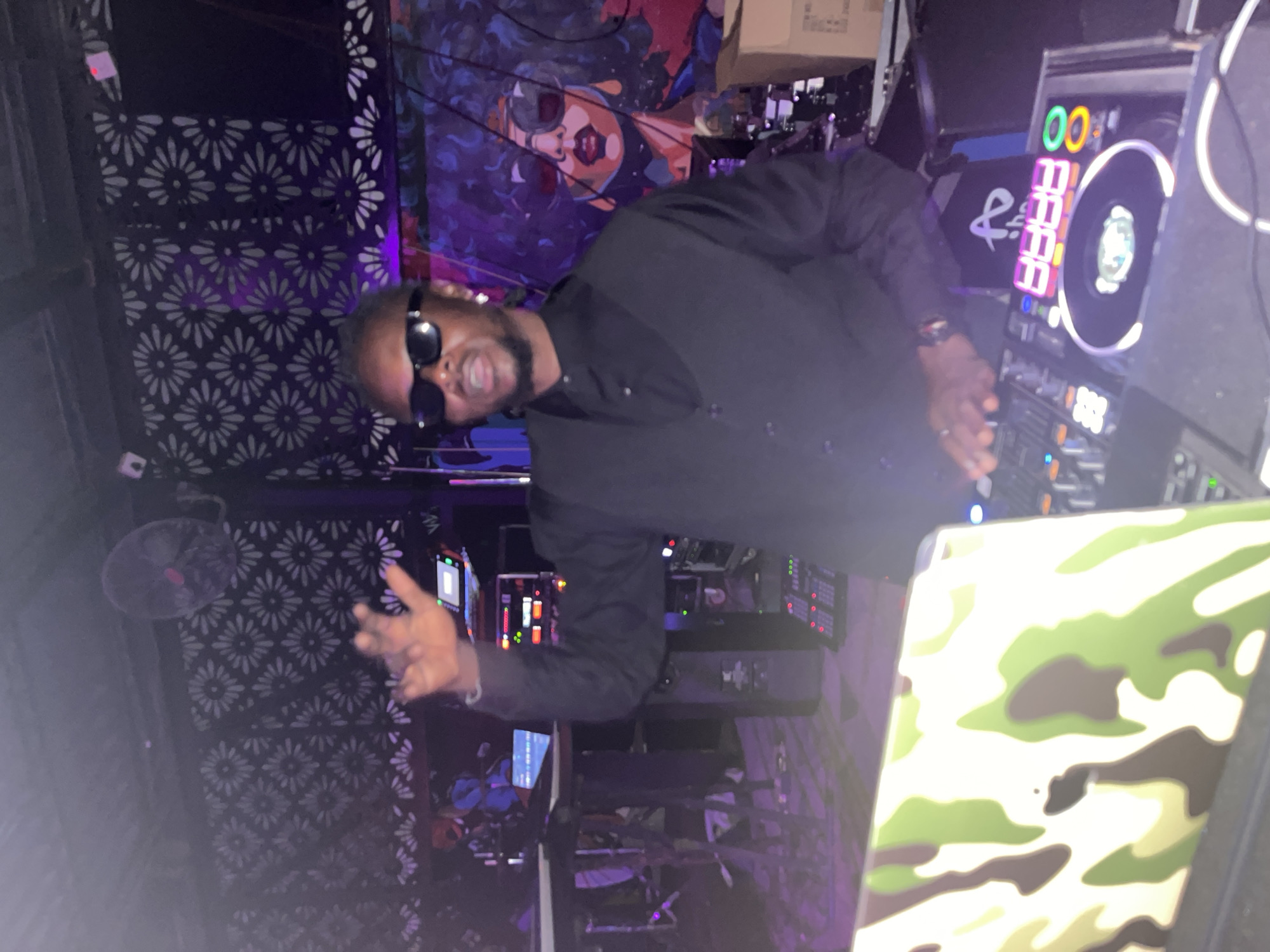 A Black male DJ, wearing a black vest over a black button-down shirt and sunglasses, stands behind a DJ setup, raising his right hand as if to hype up a crowd. He is positioned in front of a backdrop featuring a floral pattern and purple lighting. To his right, a vibrant mural depicts a Black woman with large sunglasses and an afro. The DJ setup includes a laptop with a camouflage cover, various electronic music equipment, and illuminated buttons and displays. The scene appears to be a nightclub or event space in Howayi, Nigeria.