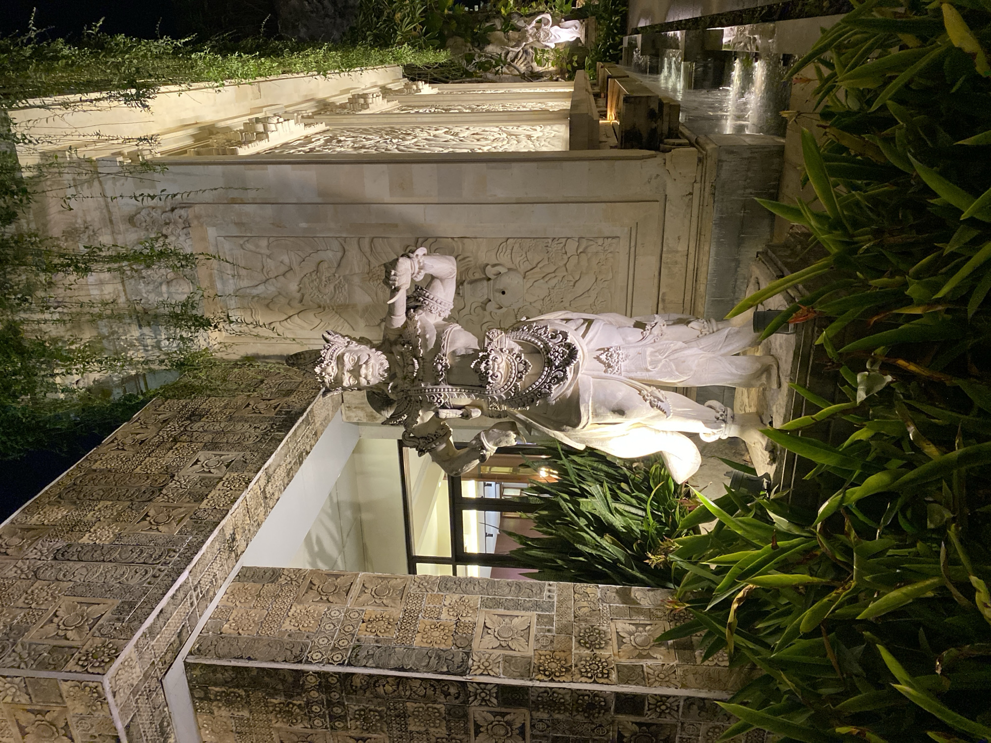 The image depicts a night scene in Kuta Selatan, Indonesia, focusing on an ornate stone statue of a Balinese figure, possibly a deity or guardian, positioned outdoors. The statue is illuminated by artificial lighting, casting shadows and highlighting its intricate details. The figure is adorned with elaborate headdresses and jewelry, and holds an object that resembles a horn or flute.
The statue stands amidst lush green foliage, with tropical plants in the foreground. Behind the statue, a building features detailed stone carvings and decorative motifs, typical of Balinese architecture. Hanging vines cascade down the building's facade, adding to the natural ambiance. Adjacent to the statue is a water feature with cascading water, reflecting the ambient light. In the background, another smaller statue is faintly visible, also illuminated. The overall impression is one of a culturally rich and tranquil setting, possibly a temple, resort, or public space in Bali.