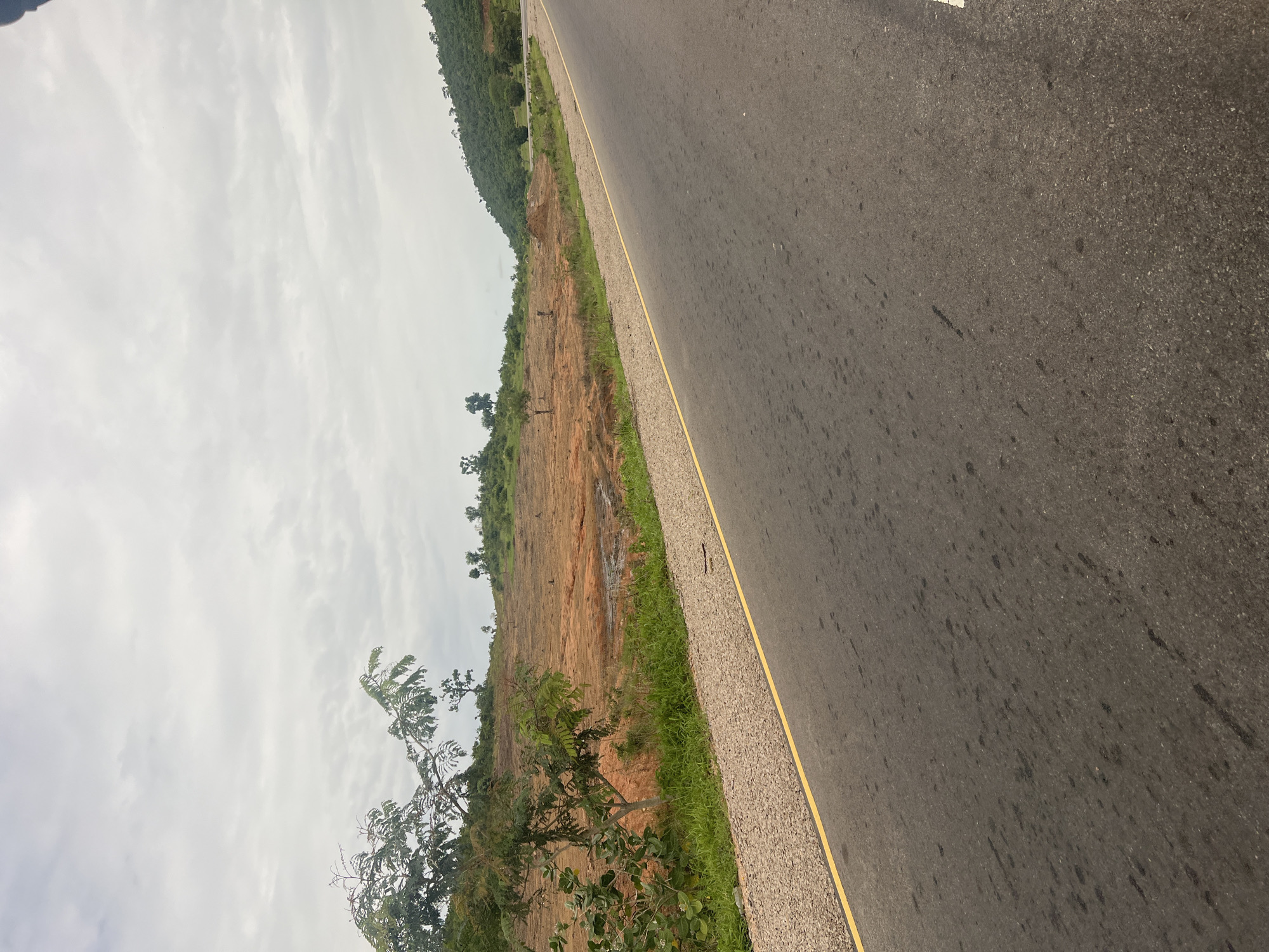 The image depicts a paved road running diagonally through a landscape. The road surface is dark asphalt, with a distinct yellow line marking the edge closest to the viewer. To the left of the road, there is a strip of gravel, followed by a red, earthen hillside dotted with sparse vegetation and some trees. Further to the left, the hillside rises into a densely wooded area, with a green, tree-covered mountain in the background. The sky is overcast with scattered clouds, suggesting a muted light and possibly a day without direct sunlight. The scene appears to be a rural or semi-rural setting in Minda, Nigeria, given the provided location context. There are no people or specific objects that stand out, beyond the natural elements and the road infrastructure. The overall impression is one of a journey or transit through a natural, somewhat undeveloped area.