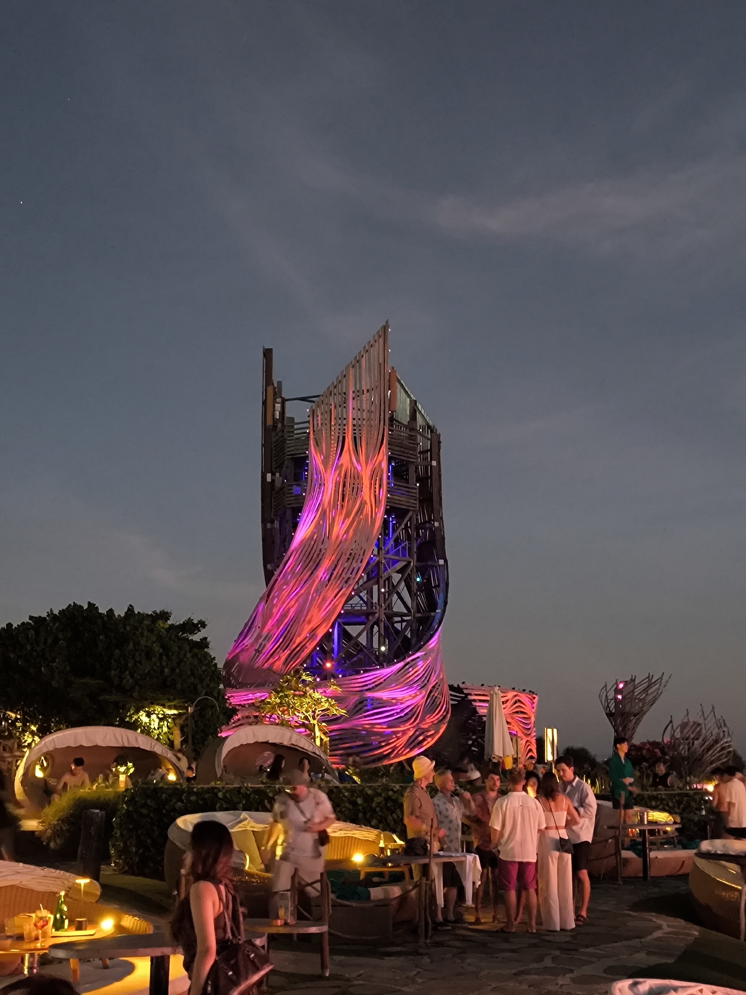 The image captures a vibrant night scene at a venue with an architectural structure, likely a tower, illuminated with pink, red, and blue lights. The structure is a prominent feature against the darkening sky. Below the tower, groups of people are gathered, some standing and conversing, others seated in what appear to be lounge areas with canopies. There are illuminated tables with drinks, suggesting a bar or restaurant setting. The environment suggests an outdoor venue with a lively social atmosphere. The location is identified as Kediri, Indonesia.