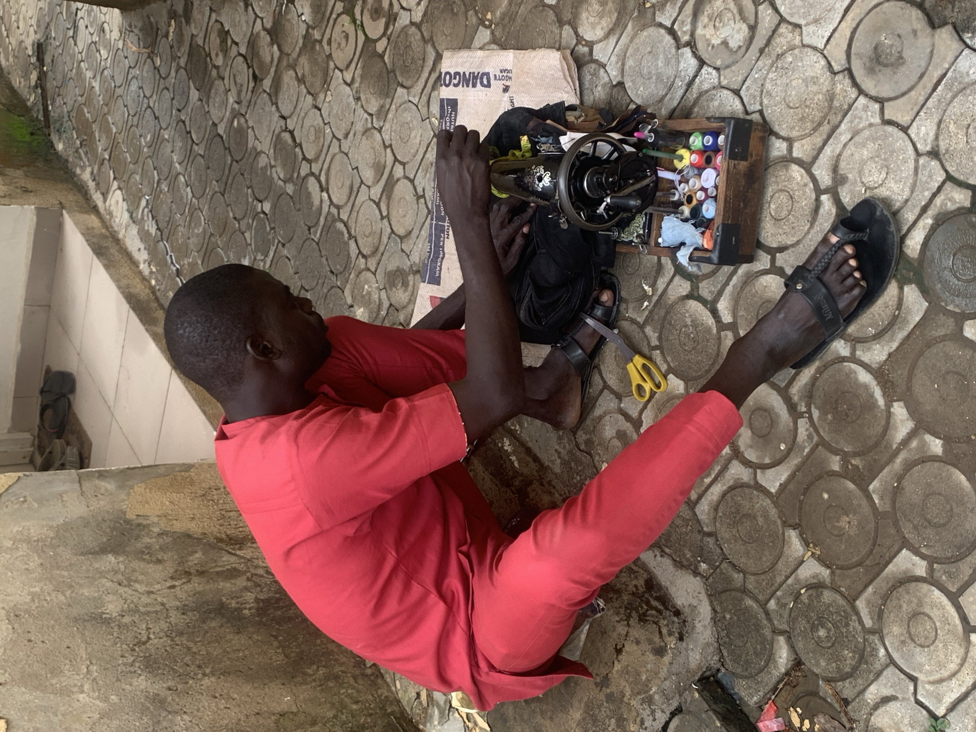 A man in a red, matching shirt and pants is sitting cross-legged on a patterned concrete surface. He is operating a black, vintage sewing machine placed on a cardboard box labeled "DANGOT" which likely refers to Dangote Cement, a common product in Nigeria. Next to the sewing machine is a wooden box filled with colorful spools of thread. A pair of yellow scissors is lying on the ground near his right foot, which is clad in a black sandal. The location is likely an urban area in Nigeria, possibly Abuja, indicated by the "DANGOT" reference and the style of the concrete surface. The man is focused on sewing, with his hands positioned to guide fabric through the machine.
