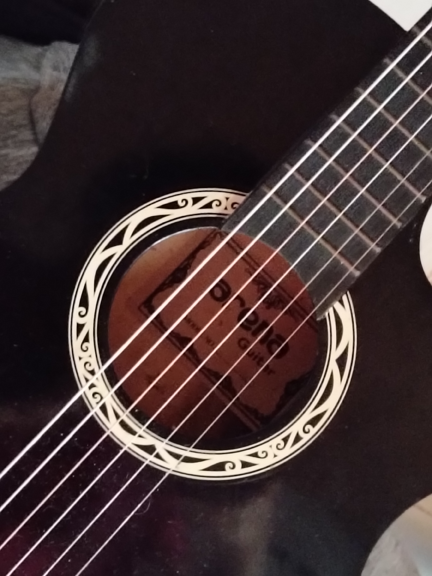 The image shows a close-up, overhead view of a black acoustic guitar. The guitar features a soundhole with a decorative rosette, and six nylon strings are visible. The fretboard and part of the headstock are also in view, angled across the body of the guitar. A label is visible inside the soundhole, partially obscured by the strings, with text that appears to read "Morena Guitar". The guitar is lying on a dark, textured surface, possibly a couch or bed, with a hint of grey fabric in the upper left corner. There are no people or any other identifying features of a specific location in the image. The image does not provide any information that would allow for its location to be determined within Davao City, Philippines, or any other city.