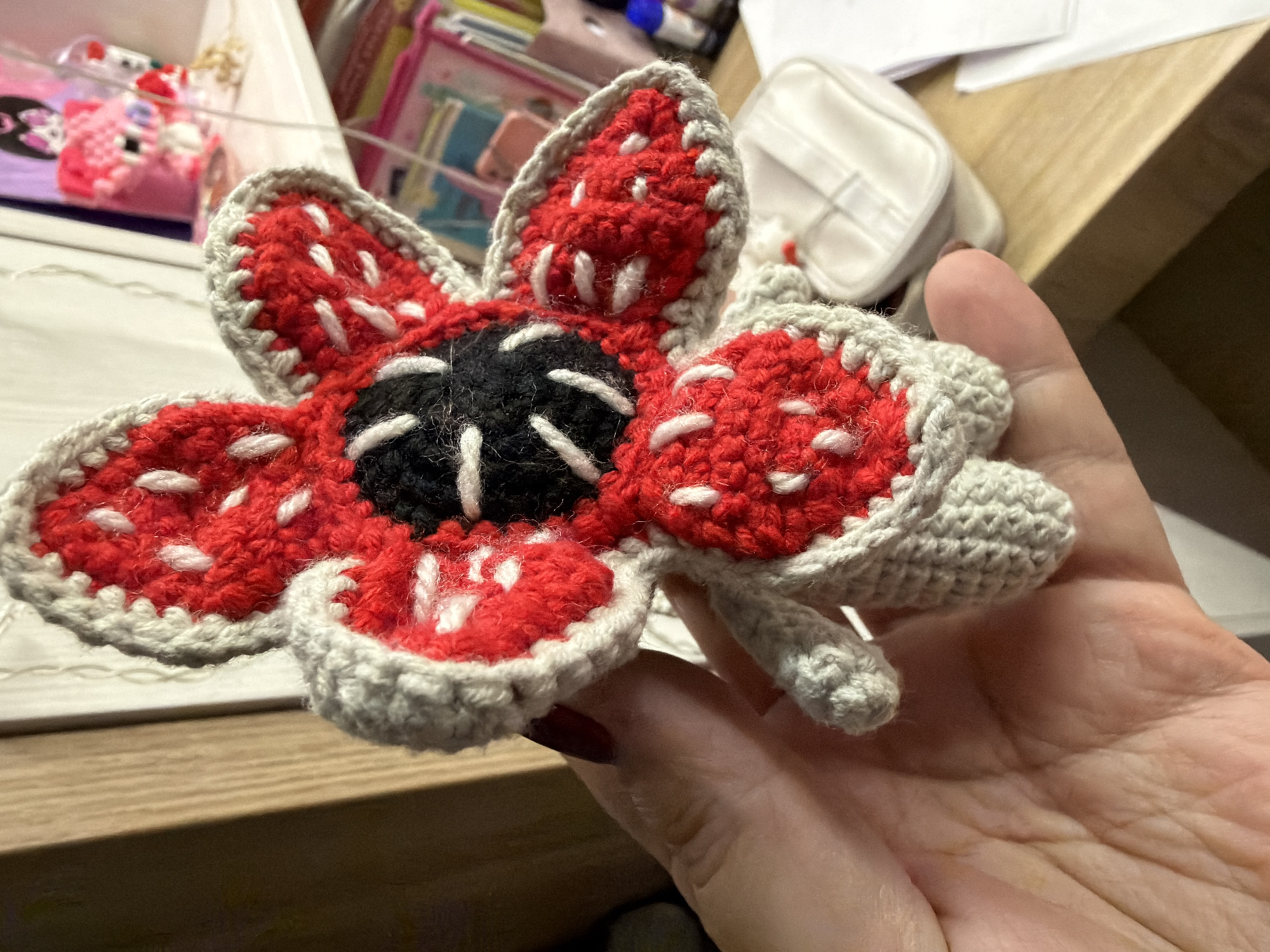 A person's hand holds a crocheted Demogorgon flower (from the "Stranger Things" TV show). The flower has a black center with white stitch-like details, surrounded by red petals with white stitch-like details and a grey crocheted border. The person's hand has maroon nail polish on one visible fingernail. In the background is a cluttered desk with a white container, books, and other miscellaneous items. The location is a room with a wooden desk. There is no information about the city of Chaikovsky, Russia.