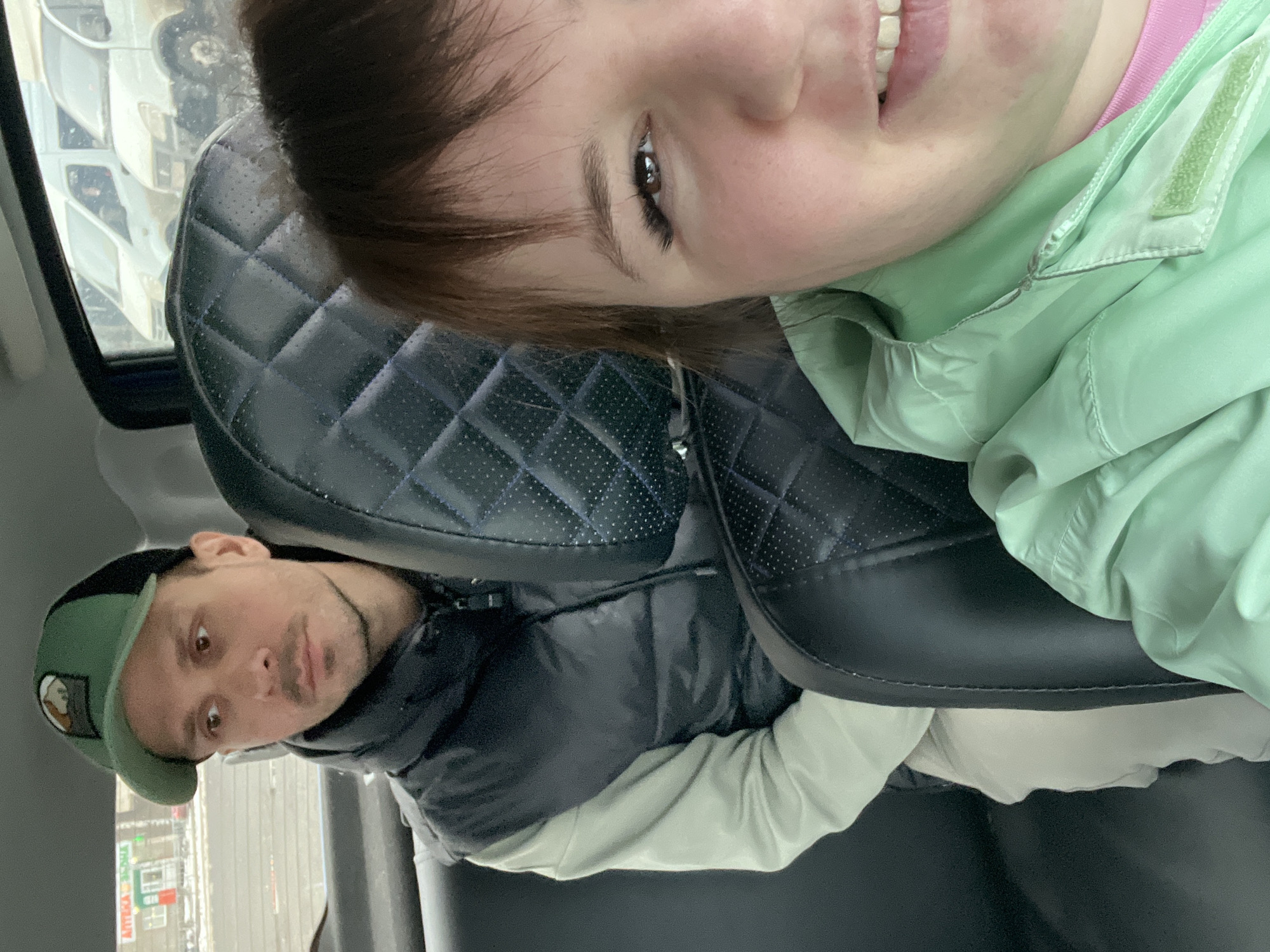 Two people are inside a vehicle. A woman is seated facing the camera, wearing a light green jacket over a pink shirt. A man is seated behind her and to the left, wearing a dark puffer jacket and a green cap with a patch. The car seats are black leather. Outside the vehicle's window is a building with signage in Russian, including the word "АПТЕКА" (pharmacy). The building appears to be in Izhevsk, Russia, based on the Cyrillic script on the signage.