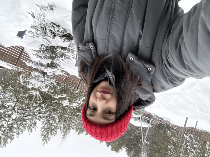 A female subject is captured in a selfie-style photograph, standing outdoors in a snowy, wooded environment identified as Sysert, Russia. The subject, positioned centrally and slightly to the right, wears a bright red knit beanie and a dark gray, quilted winter coat with a prominent collar. Her dark hair is visible beneath the beanie, extending to her shoulders. She looks directly towards the camera with a neutral expression.
The background is dominated by snow-covered terrain, indicating a winter setting. To the left and behind the subject, a snow-covered incline features faint tracks. Further back on the left, two white vehicles are partially visible, parked near a building with a light-colored roof. A brown slatted fence runs horizontally across the midground, separating the immediate area from denser forest. Multiple snow-laden pine and fir trees are present, both directly behind the subject and throughout the background, suggesting a forest or park area. A small, snow-covered coniferous tree is visible to the subject's right. The sky is overcast and white.