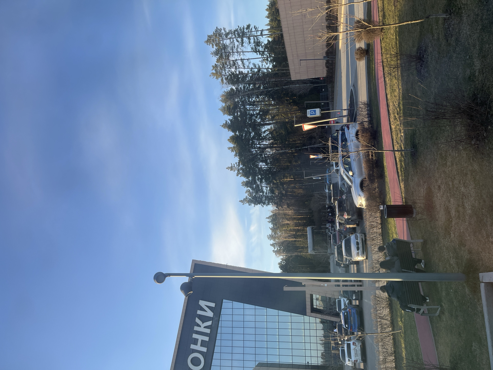 The image depicts an outdoor scene, likely a parking area near a building, possibly located in Истра Гоу Доц, Russia. The sky is blue with light clouds.
Several cars are parked in rows. A building with a modern architectural design is visible, featuring a glass facade. A sign with the word "ОНКИ" (likely part of a larger sign) is attached to the building.
Trees line part of the parking area. A pedestrian pathway, partially painted red, runs alongside the parking area.
A bench sits on a grassy area, with a person sitting on it.