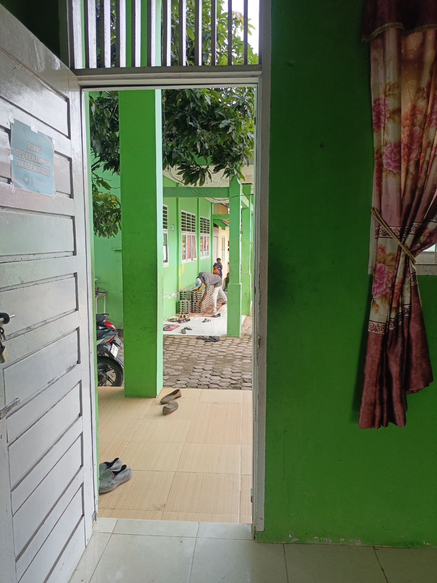 The image depicts an outdoor hallway or walkway with green painted pillars and walls. In the foreground, a white door is ajar, revealing a tiled floor with a pair of grey slip-on shoes. Beyond the door, the hallway continues with a tiled floor and a patterned walkway. Several pairs of sandals are scattered on the walkway.
A person wearing a grey long-sleeved shirt and light-colored pants is bent over, appearing to arrange items on the patterned walkway. To the left, a black motorcycle is parked. Above, green foliage from trees overhangs the hallway. In the background, a building with white-framed windows is visible. On the wall to the right, a floral patterned curtain is tied back with a rope. A sign on the white door reads "RUANG TES KEMAMPUAN AKADEMIK 2026" and "TEN NEGRI 18 BANDASAK". This suggests the location is a classroom or testing room at "Negeri 18 Banda Sak" in Lhokseumawe, Indonesia, likely for an academic ability test in 2026. The overall impression is one of an institutional setting, possibly a school or public building, during daytime.