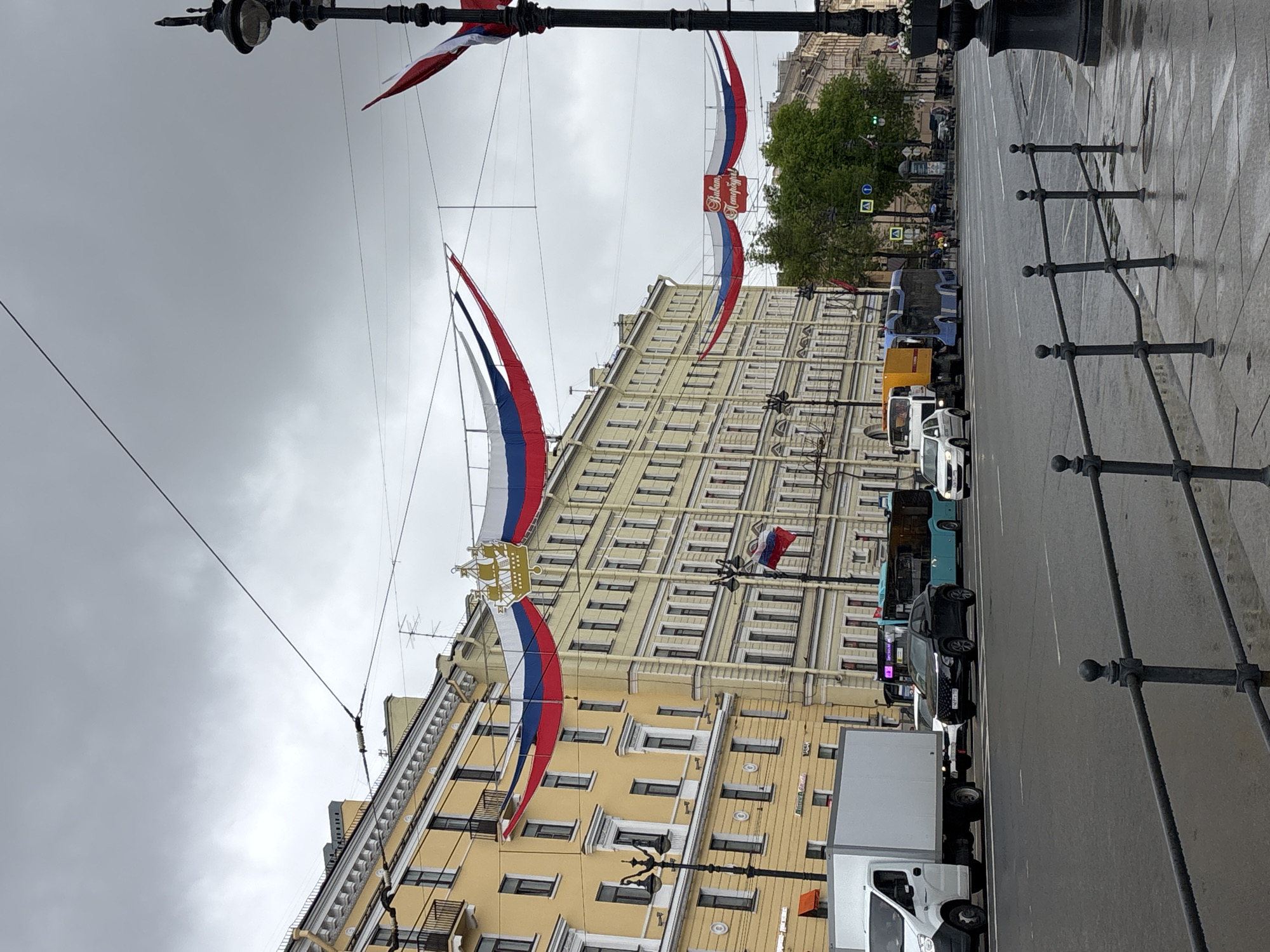 The image depicts a street in Saint Petersburg, Russia, under a cloudy sky. The street is lined with buildings, predominantly yellow in color, adorned with Russian flags and banners strung across the street. A variety of vehicles, including cars, trucks, and buses, are visible along the street. The pavement appears wet, suggesting recent rain. Black, ornamental iron fencing runs along the edge of the sidewalk. A street lamp is in the foreground, framing the scene.