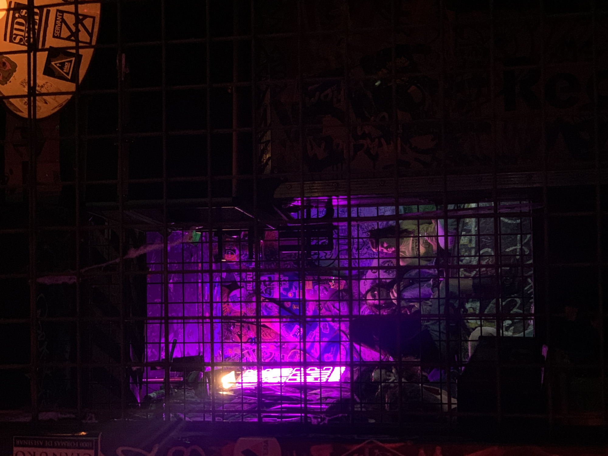 The image depicts a dark, urban nighttime scene viewed through a prominent metal grid, likely a security gate or window covering. The primary light source emanates from within the structure behind the grid, casting an intense, neon purple glow.
In the center of the frame, behind the grid and bathed in purple light, is a cluttered interior space. Various shapes, possibly posters, graffiti, or abstract art, are visible on the walls. A bright, warm, orange-yellow light, possibly a bare bulb or lamp, is positioned on the left side of this purple-lit area, creating a stark contrast. On the right, a figure with what appears to be blonde hair can be faintly seen looking downwards, possibly a mannequin or a person inside.
In the upper left corner, outside the main purple-lit section, a dim orange light illuminates several stickers and pieces of paper. Clearly visible text includes "SID'S" and "SERVANT," along with a black triangle logo. The upper right portion of the image is darker, but faint graffiti or markings, possibly including "KCG" or "KRO," are discernible on a wall.
At the very bottom of the frame, partially obscured by the grid, white text on a dark background reads "1000 FORMAS DE ASESINAR" (1000 ways to kill), with "GRANERO" visible below it. The overall impression is that of a closed shop, bar, or an artistic display with a distinct, somewhat edgy and enigmatic atmosphere, captured at night. No direct activity is taking place.